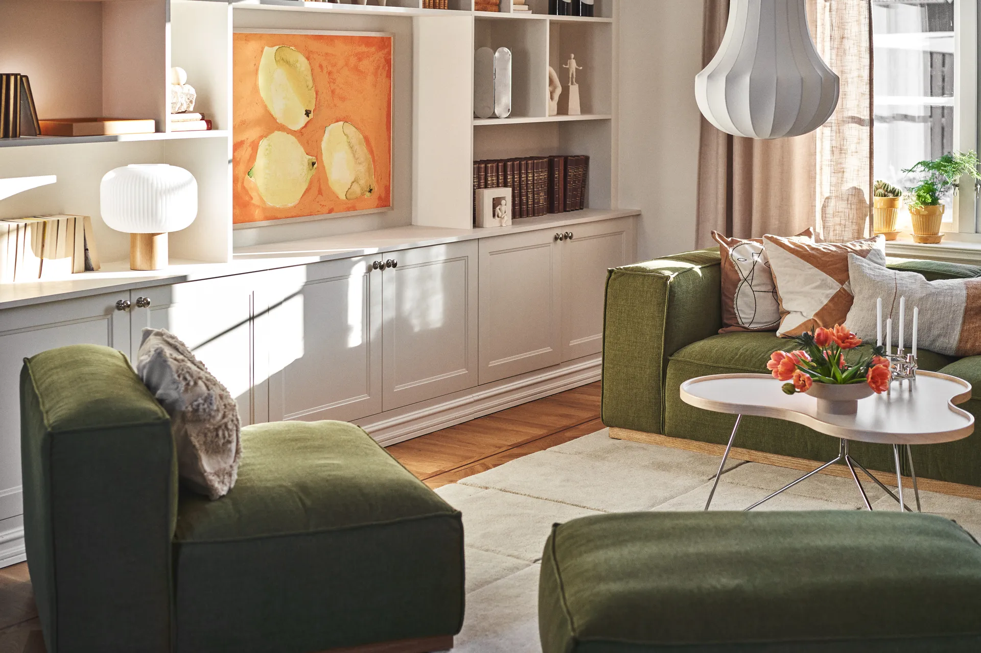 Sala de estar ensolarada com sofás verdes, uma mesa de centro branca com tulipas laranjas e velas, e prateleiras brancas com arte.