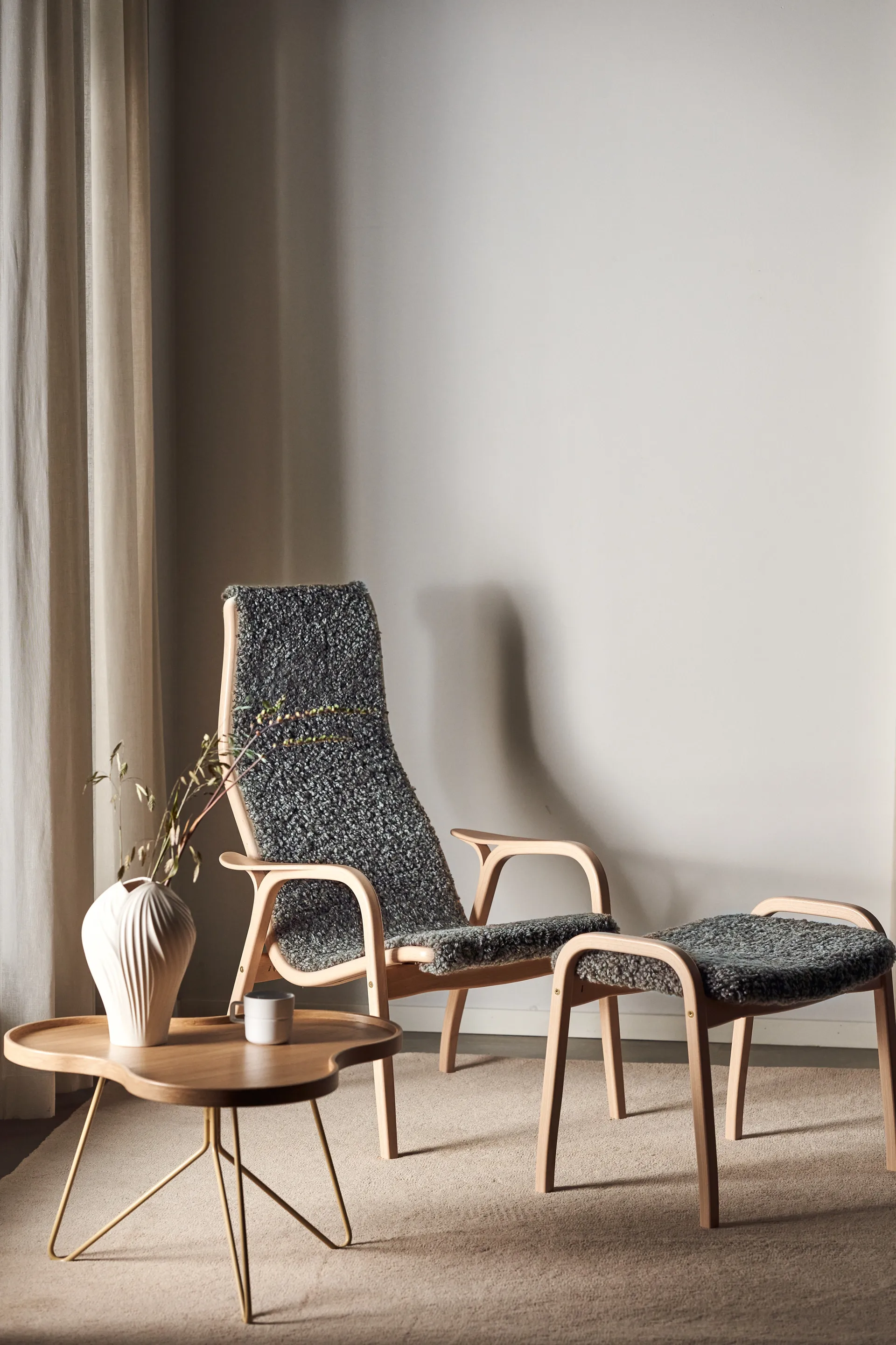 Cadeira de sala e pufe a condizer em madeira clara com estofo texturizado cinzento escuro, junto a uma pequena mesa de centro de madeira com pés dourados, jarra e caneca.