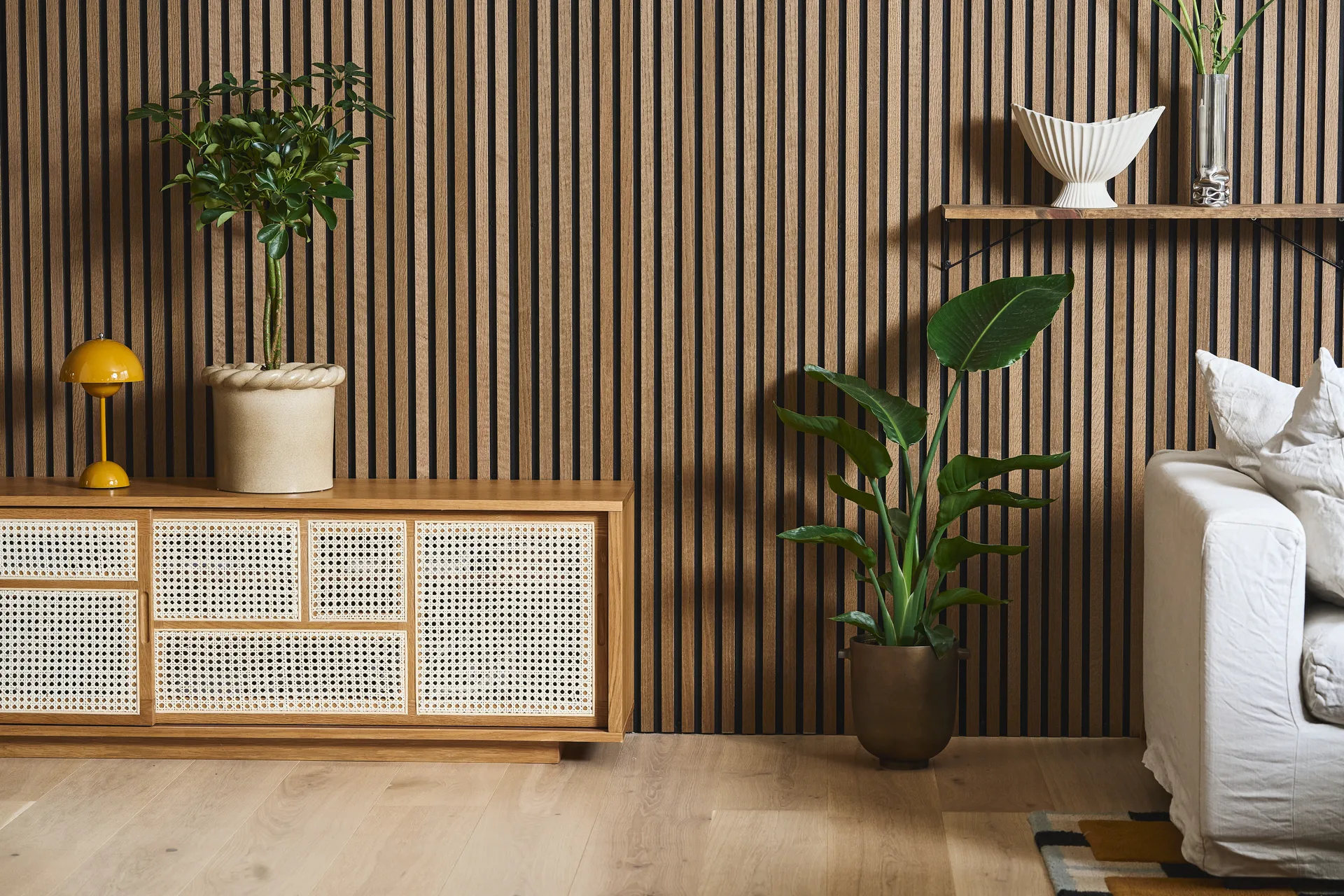 Sala moderna com parede de ripas de madeira, aparador de rotim, candeeiro amarelo, plantas e sofá branco.