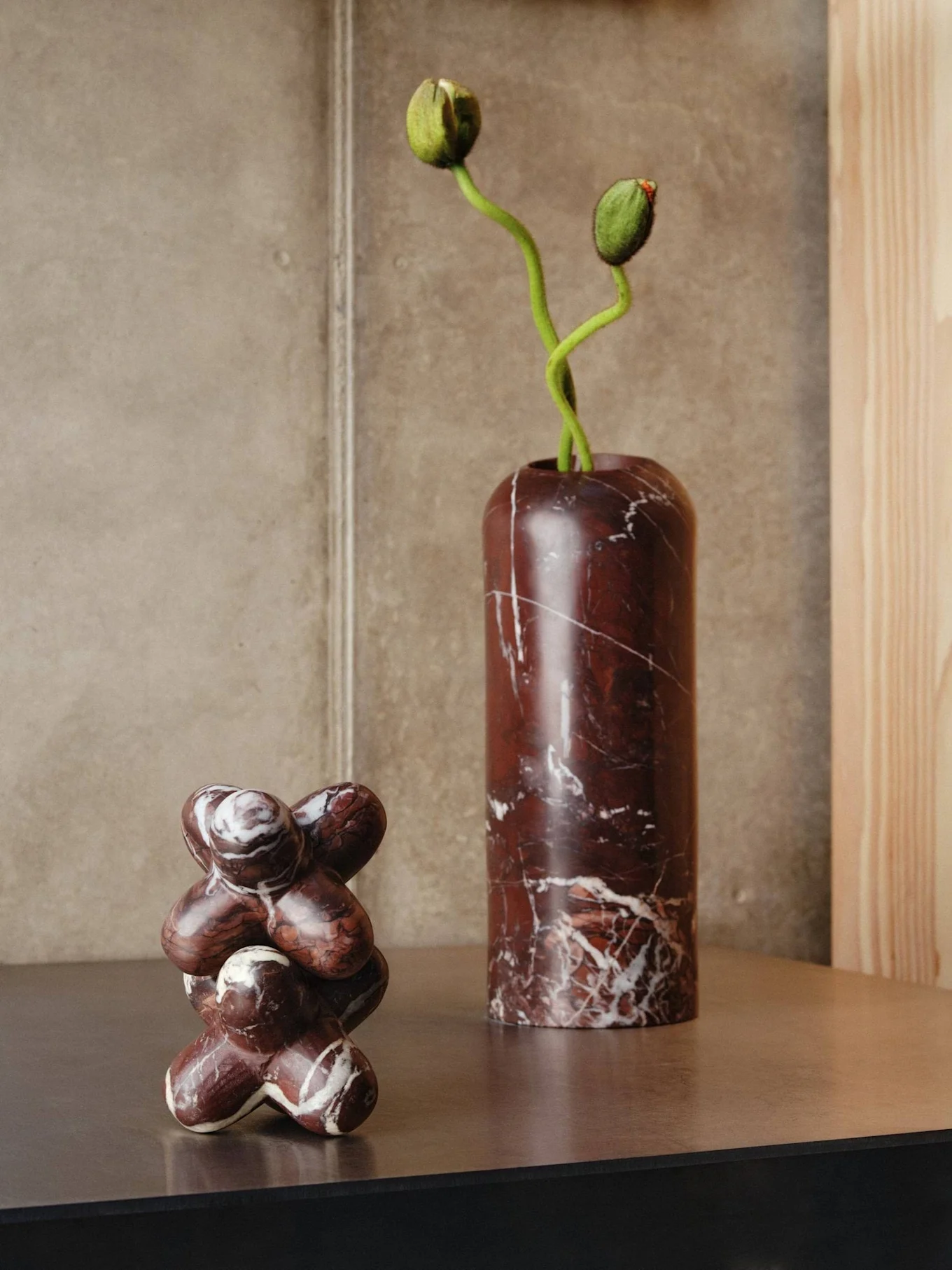 Dois objetos de mármore numa prateleira: um vaso alto vermelho escuro com botões de flores verdes e uma escultura abstrata.