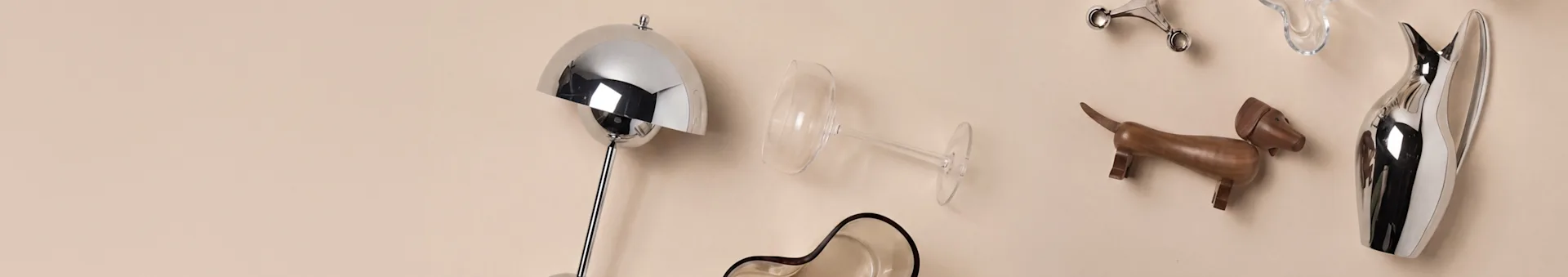 Variedade de objetos de decoração para casa, incluindo um candeeiro prateado, copo de vinho, cão de madeira e jarra prateada sobre um fundo bege.