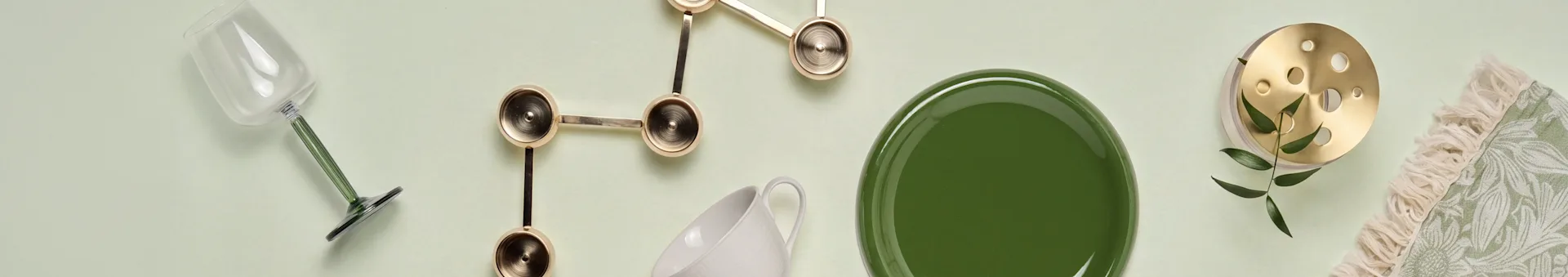 Copo com haste verde, decoração dourada, caneca branca, prato verde, objeto de latão com folhas, têxtil verde franjado.