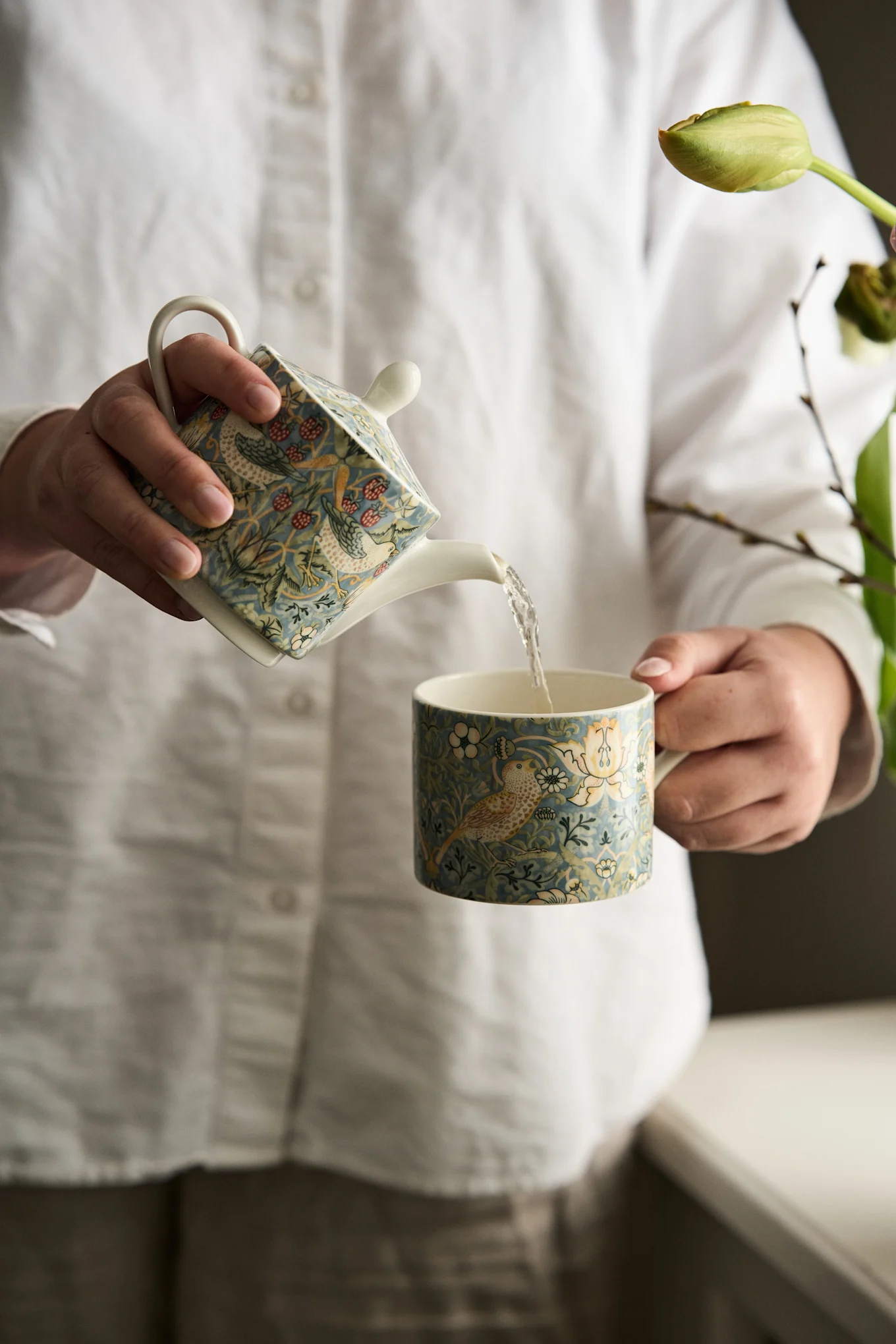 Mãos a deitar de um bule azul com padrão de pássaros e bagas para uma caneca a condizer.