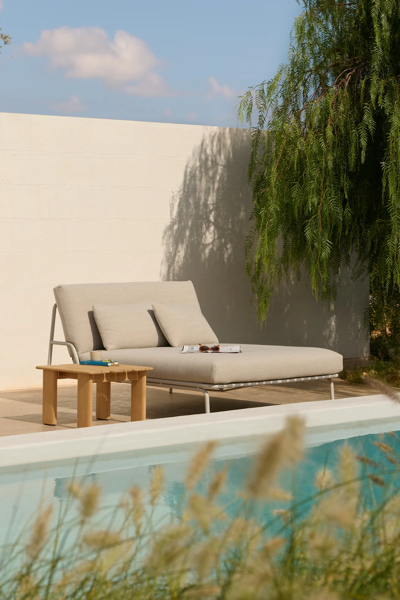 Espreguiçadeira cinza-claro com almofadas, uma mesa lateral de madeira e uma revista junto a uma piscina.