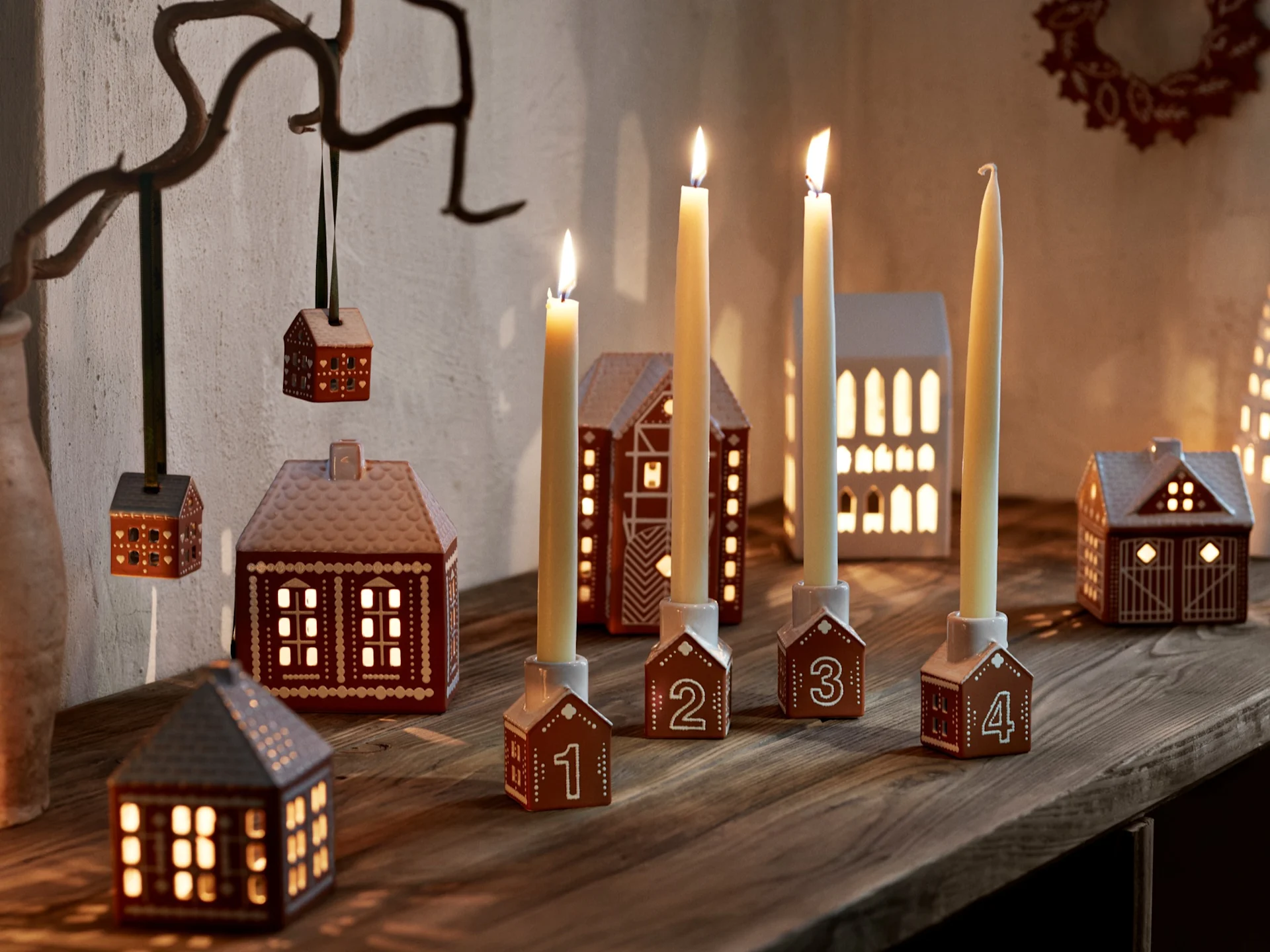 Quatro castiçais numerados em forma de casa de pão de gengibre com velas acesas e várias lanternas sobre uma superfície de madeira.