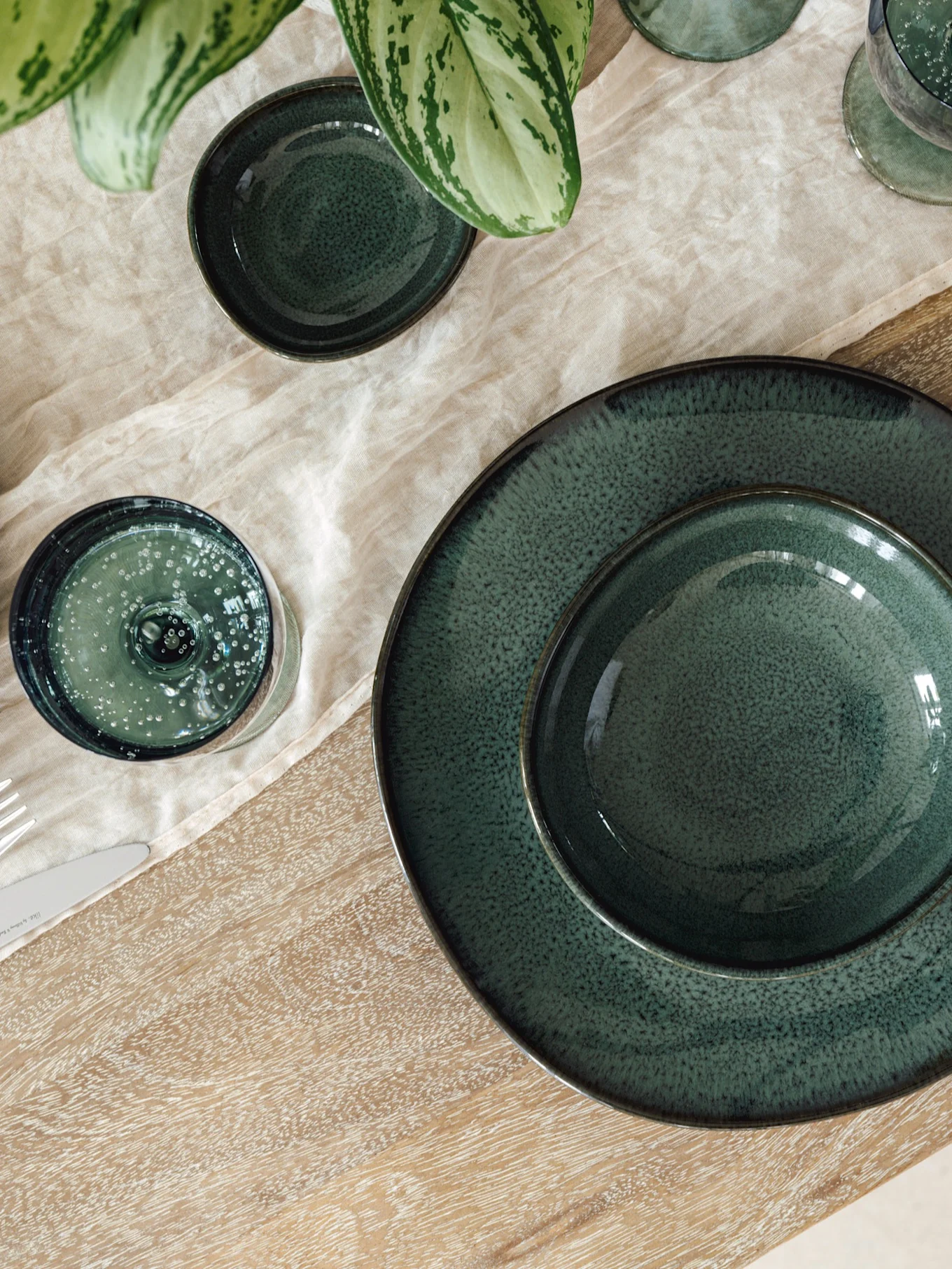 Mesa posta com pratos, taças e copos de cerâmica verde escura sobre toalha de linho claro e mesa de madeira, com planta verde.