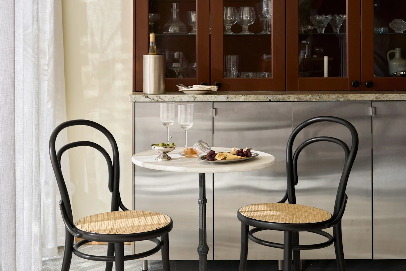Cantinho de jantar aconchegante com mesa de mármore, duas cadeiras pretas, prato de queijo, frutas, azeitonas e taças.