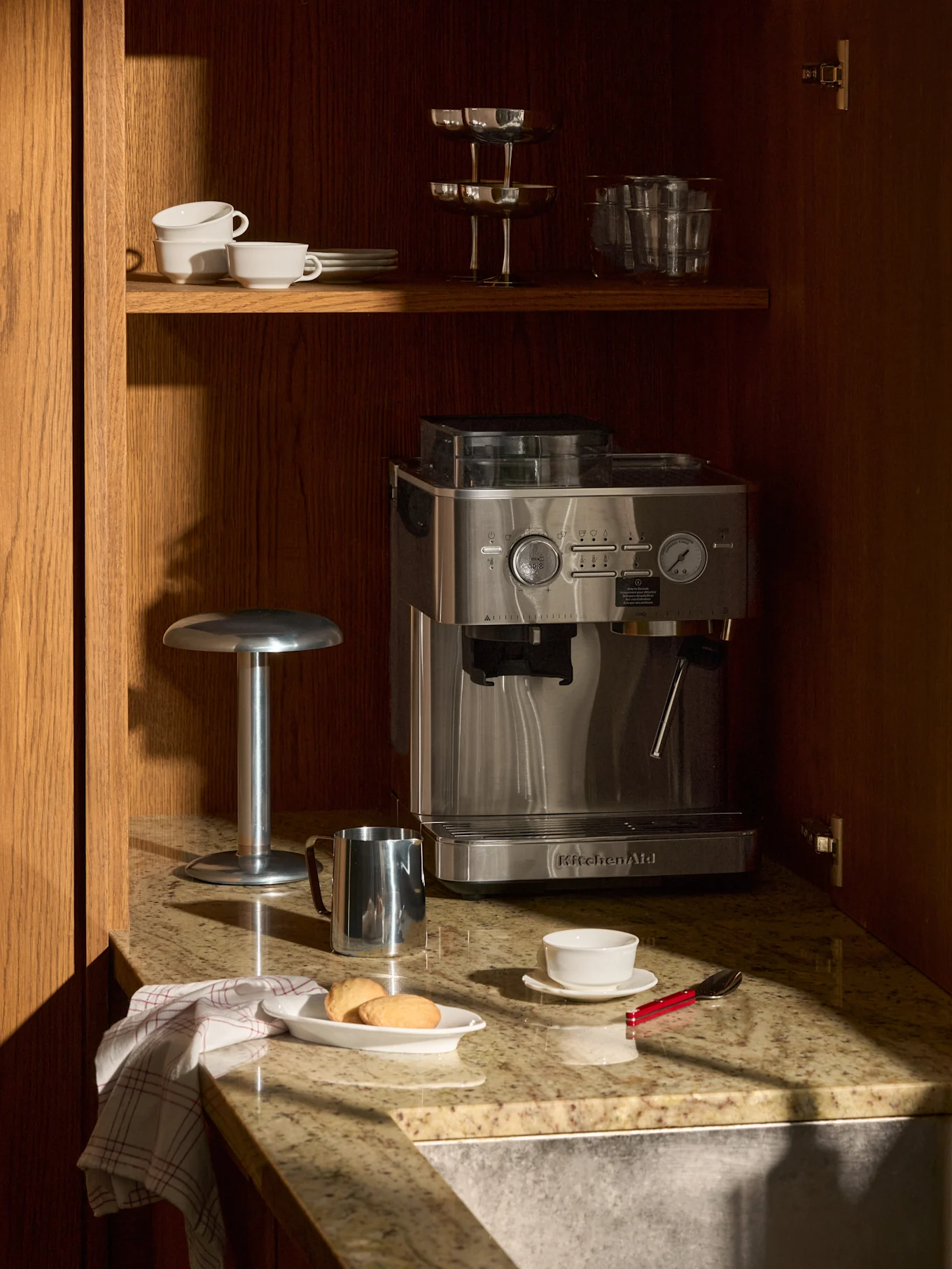 Máquina de expresso inox em balcão granito, em armário madeira. Acima, chávenas, copos. No balcão, biscoitos, chávena, jarro de leite.