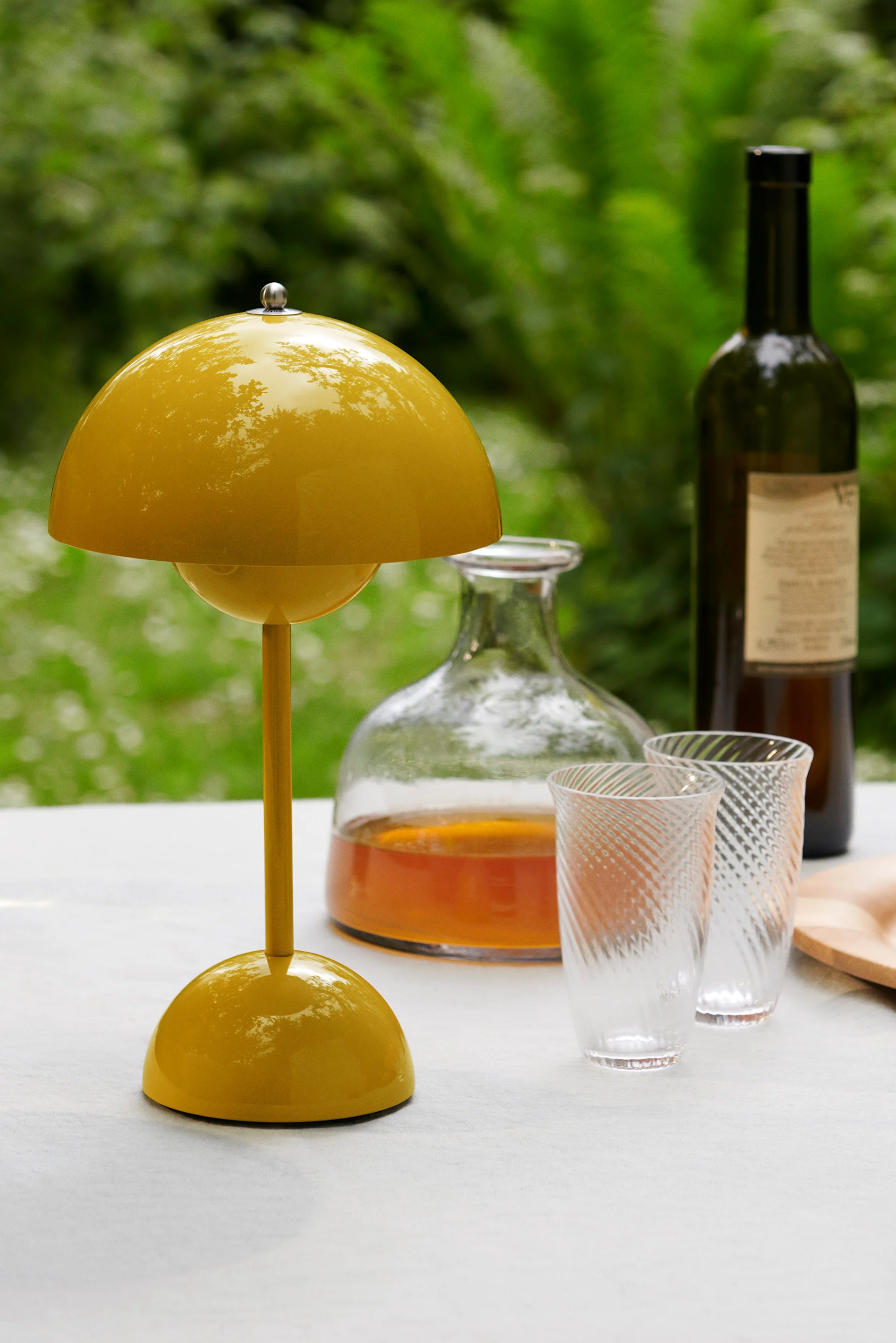 Um candeeiro de mesa amarelo brilhante em forma de cogumelo sobre uma mesa branca, com uma garrafa, dois copos com padrão e uma garrafa de vinho atrás, contra um fundo verde.