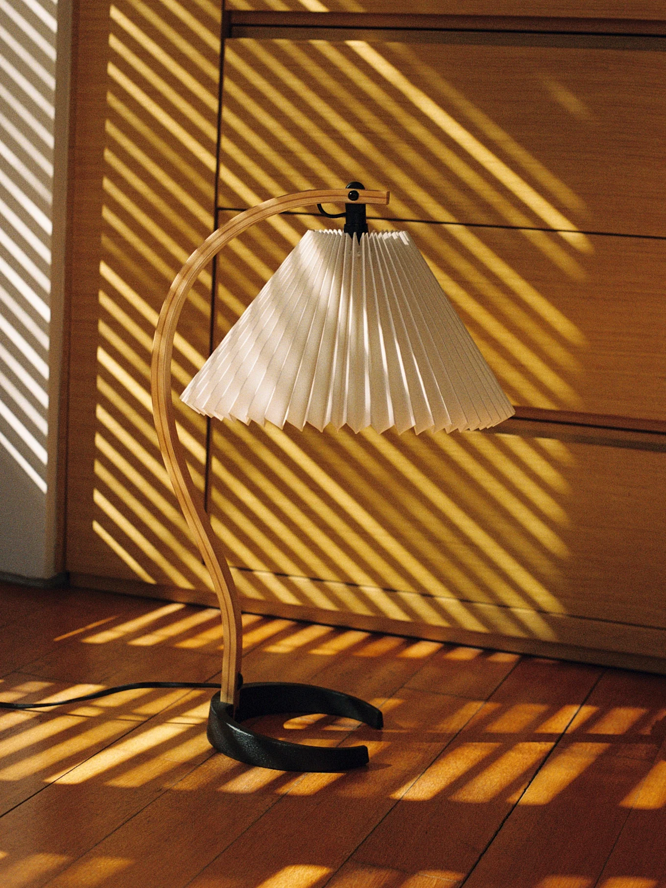 Candeeiro de secretária com suporte curvo de madeira e abajur plissado, iluminado por faixas de luz diagonais em chão de madeira.