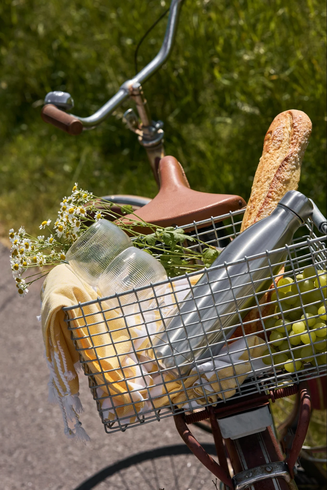 Cesto frontal de bicicleta com artigos de piquenique: uma baguete, garrafa de água, uvas, copos, toalha amarela e margaridas brancas.