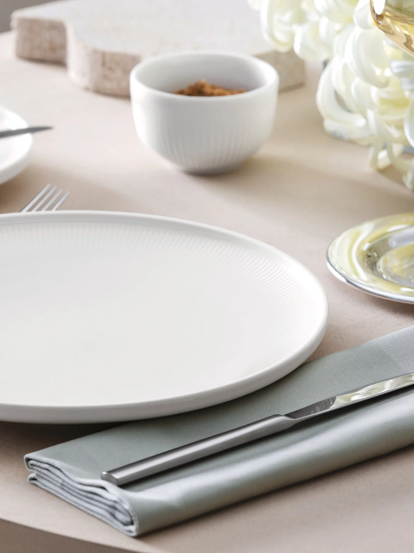 Um prato branco canelado, guardanapo verde sálvia e talheres prateados sobre uma mesa com uma pequena tigela ao fundo.