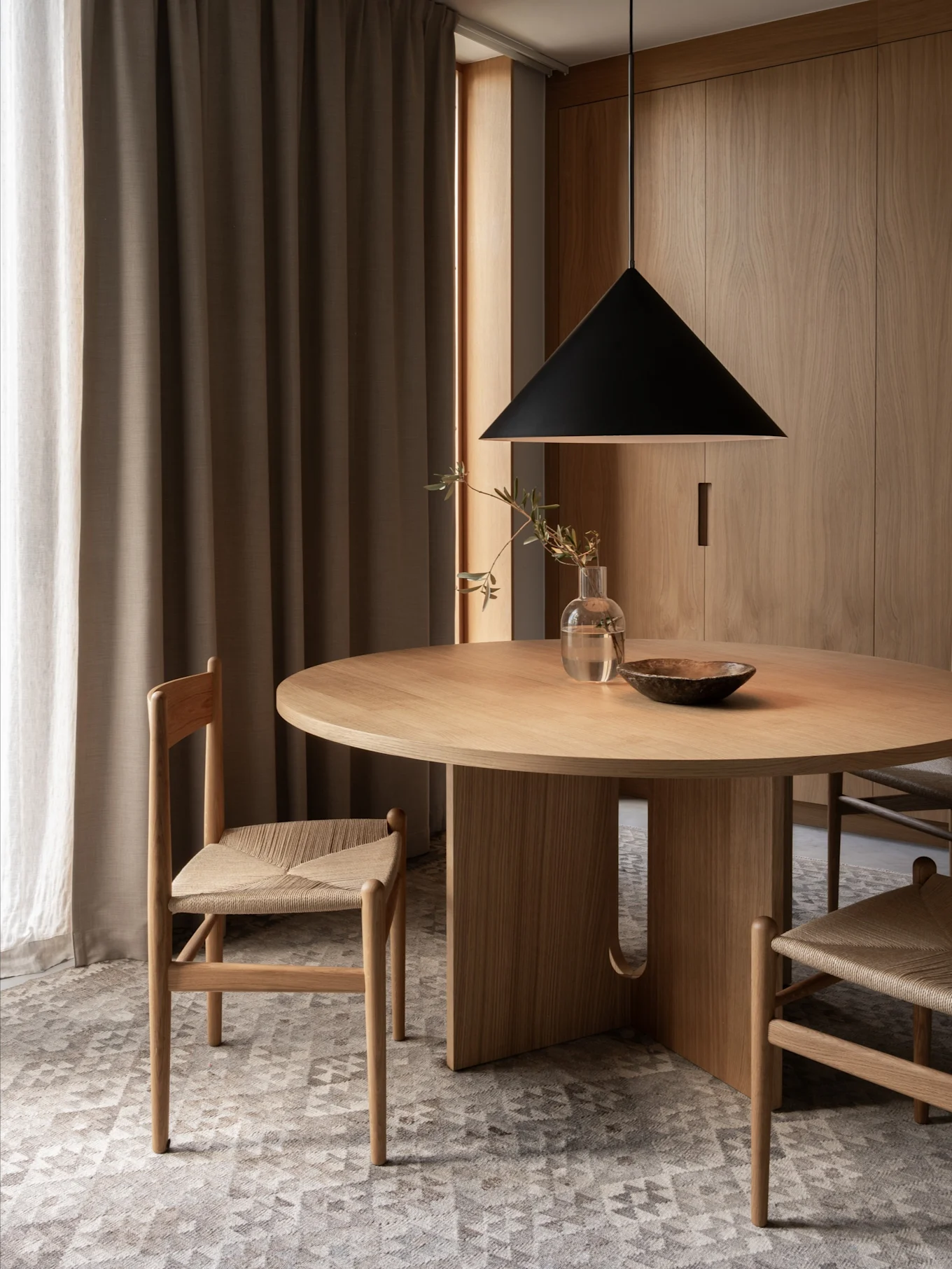 Uma área de jantar minimalista com uma mesa redonda de madeira, duas cadeiras com assentos entrançados, um candeeiro suspenso preto e paredes de madeira.