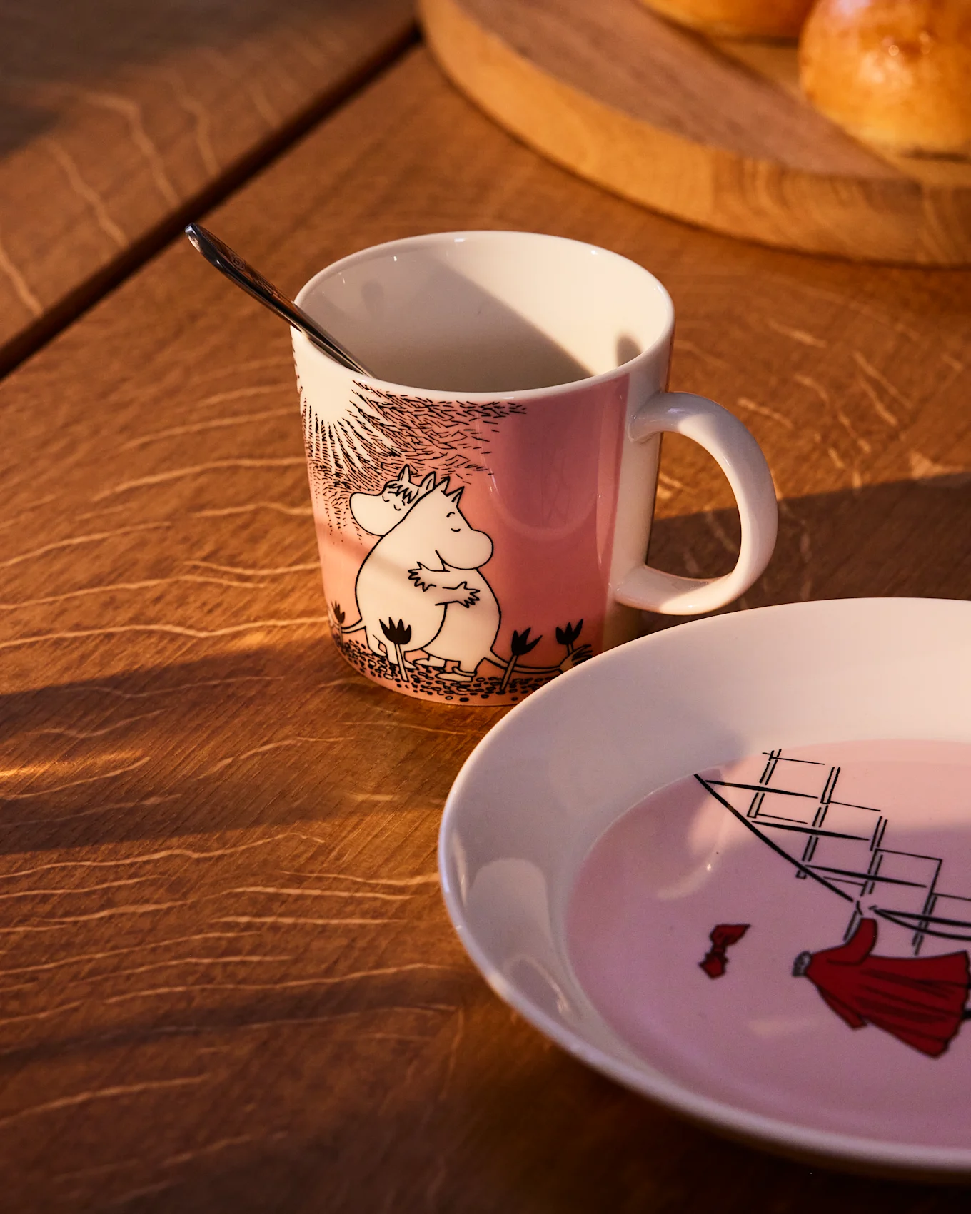 Caneca Moomin rosa com Moomintroll e Snorkmaiden, ao lado de um prato a condizer com um veleiro, numa mesa de madeira.