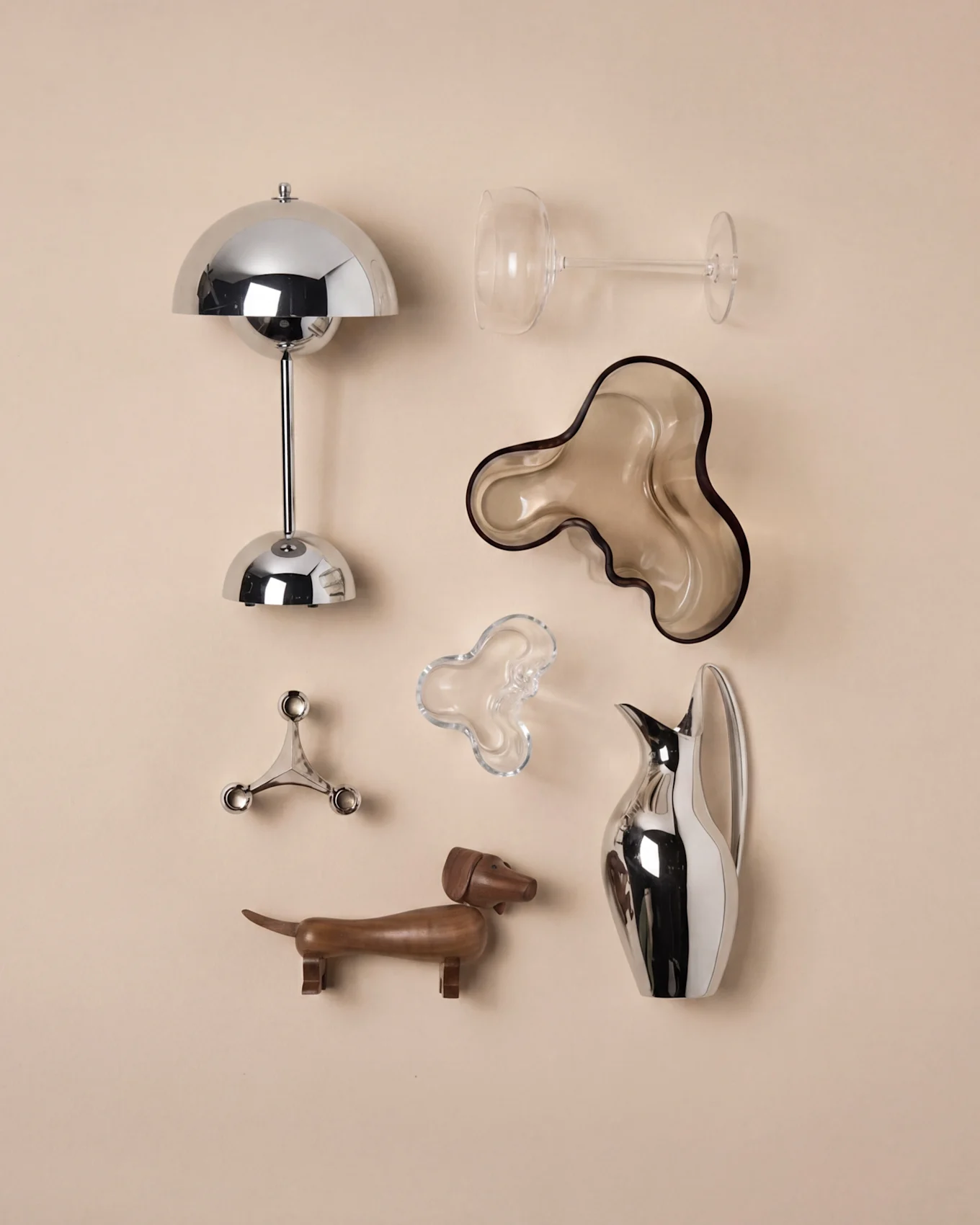 Decoração moderna: candeeiro cogumelo cromado, copo de vinho, taça ondulada castanha, castiçal prateado, dachshund de madeira, jarro prateado.