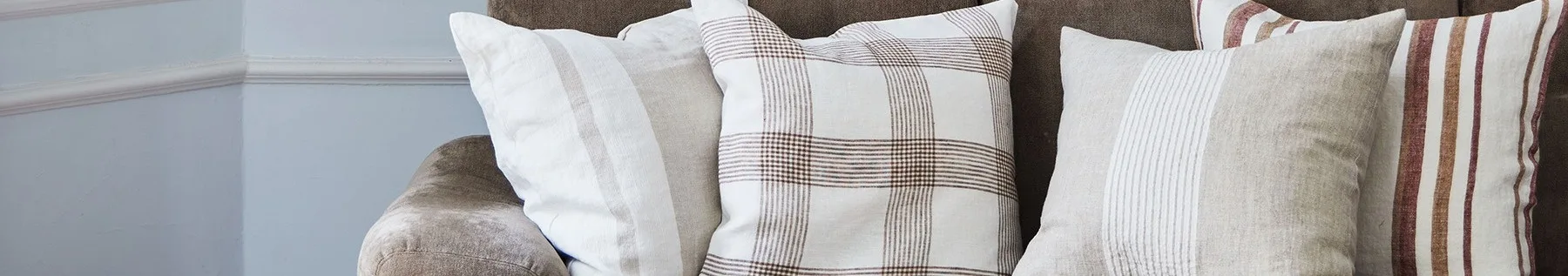 Close-up de três almofadas em um sofá marrom, apresentando padrões brancos, bege e xadrez com listras marrons e vermelhas.