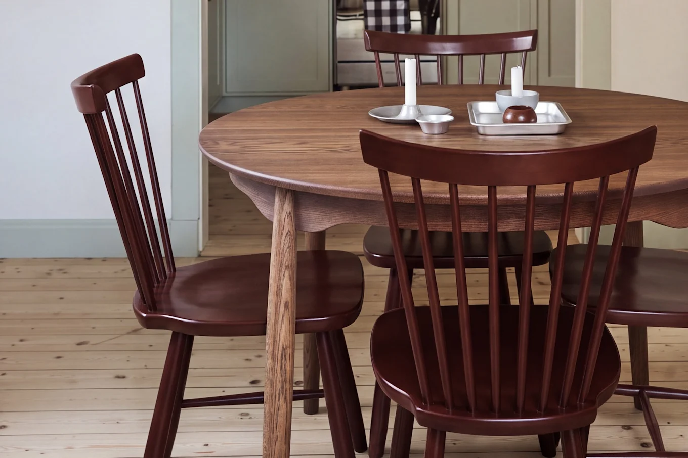 Sala de jantar com mesa redonda de madeira, cadeiras vermelhas e um candeeiro suspenso vermelho, com vista para uma cozinha com armários verdes claros.