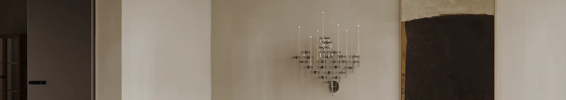 Um candelabro modular geométrico cromado moderno com várias velas brancas acesas montado numa parede clara.