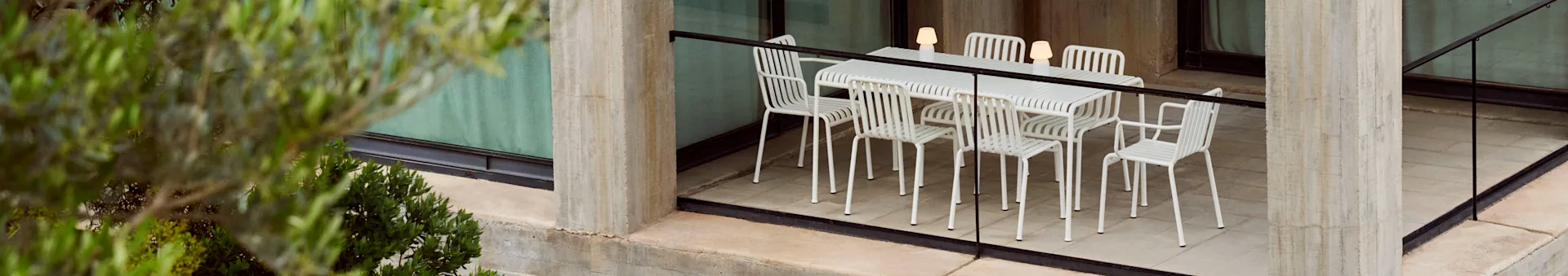 Um conjunto de jantar ao ar livre com uma mesa branca de ripas e seis cadeiras a condizer, iluminado por três candeeiros portáteis, num pátio de betão com plantas.