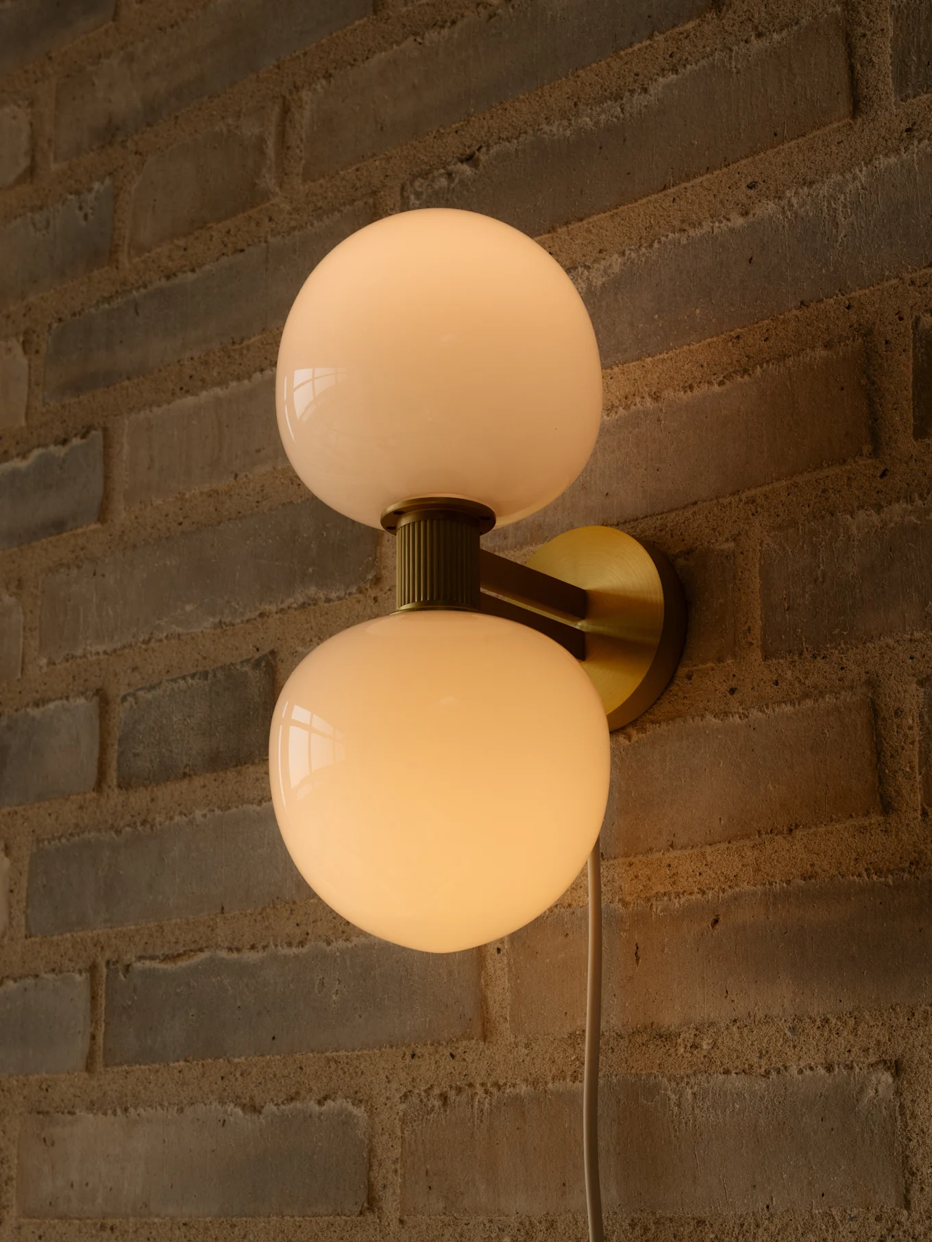 Candeeiro de parede com duas cúpulas esféricas de vidro branco leitoso e suporte de latão numa parede de tijolos.