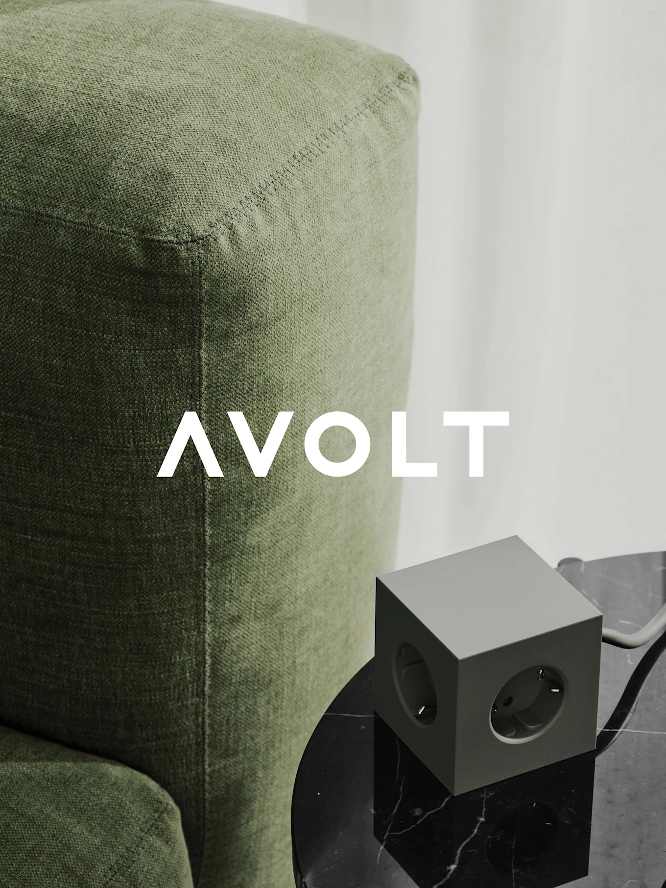 Uma tomada elétrica verde em cubo com três saídas sobre uma mesa de mármore preto junto a um sofá verde. Logótipo AVOLT.