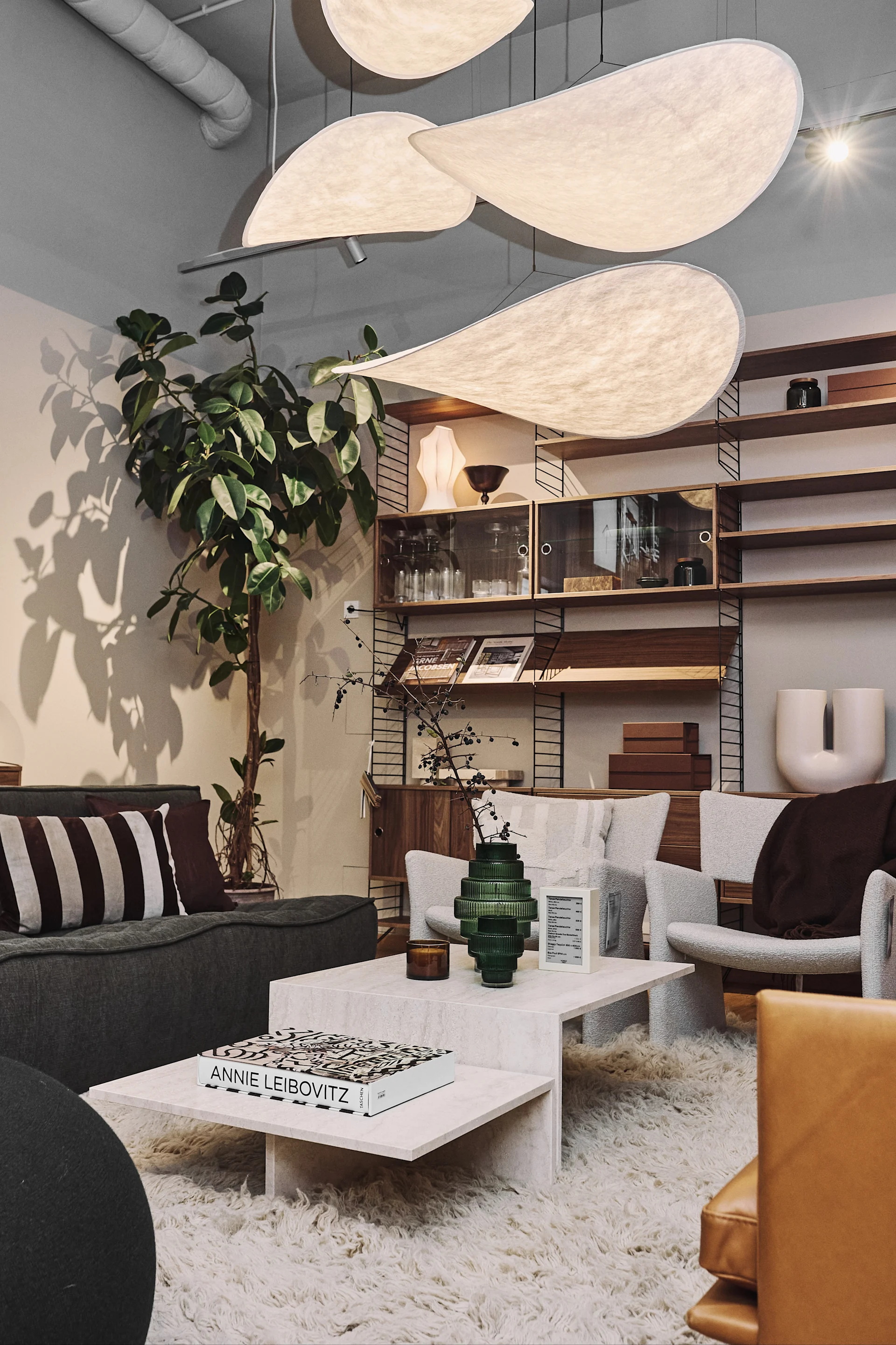 Interior com luminárias esculturais, sofá cinzento, poltronas creme, mesa de centro, tapete e prateleiras.