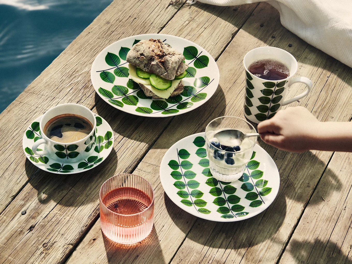 Pequeno-almoço em cais de madeira: pratos com folhas, sanduíche, café, chá, iogurte com mirtilos e bebida rosa.