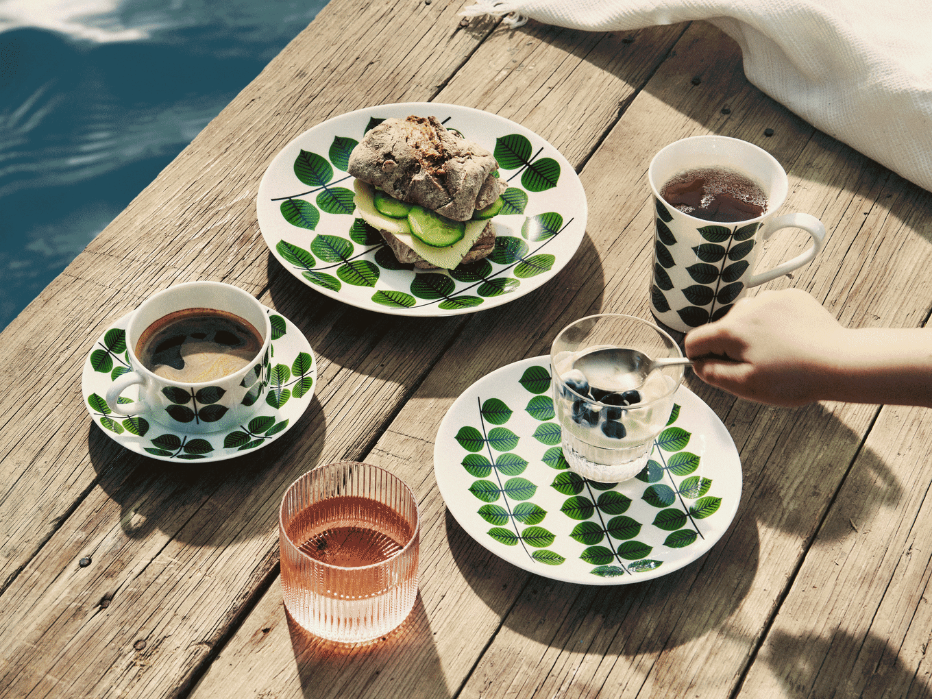 Pequeno-almoço em cais de madeira: pratos com folhas, sanduíche, café, chá, iogurte com mirtilos e bebida rosa.