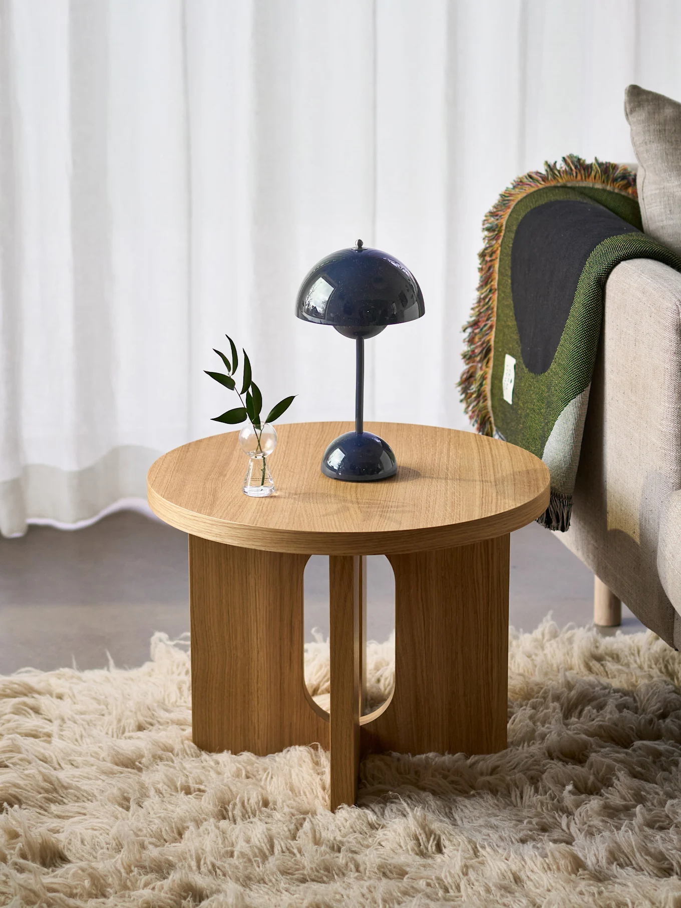 Mesa lateral redonda de madeira com abajur azul-escuro e planta, ao lado de poltrona com manta verde, sobre tapete felpudo.