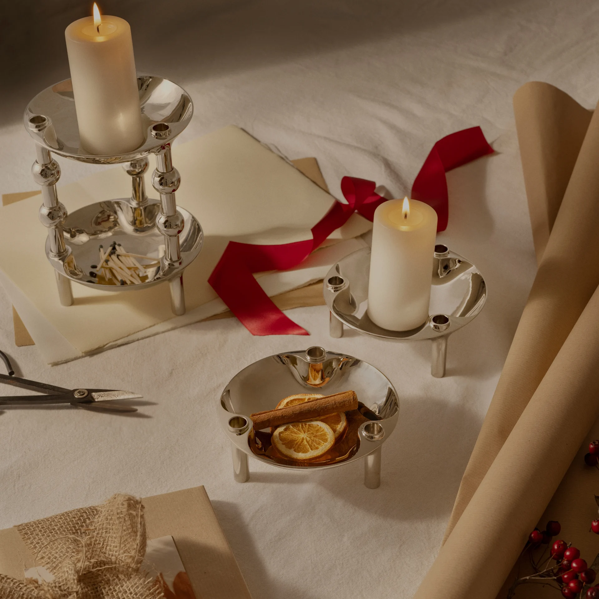 Três castiçais de prata, dois com velas acesas, um com laranjas secas e canela, dispostos com papel de embrulho e fita.
