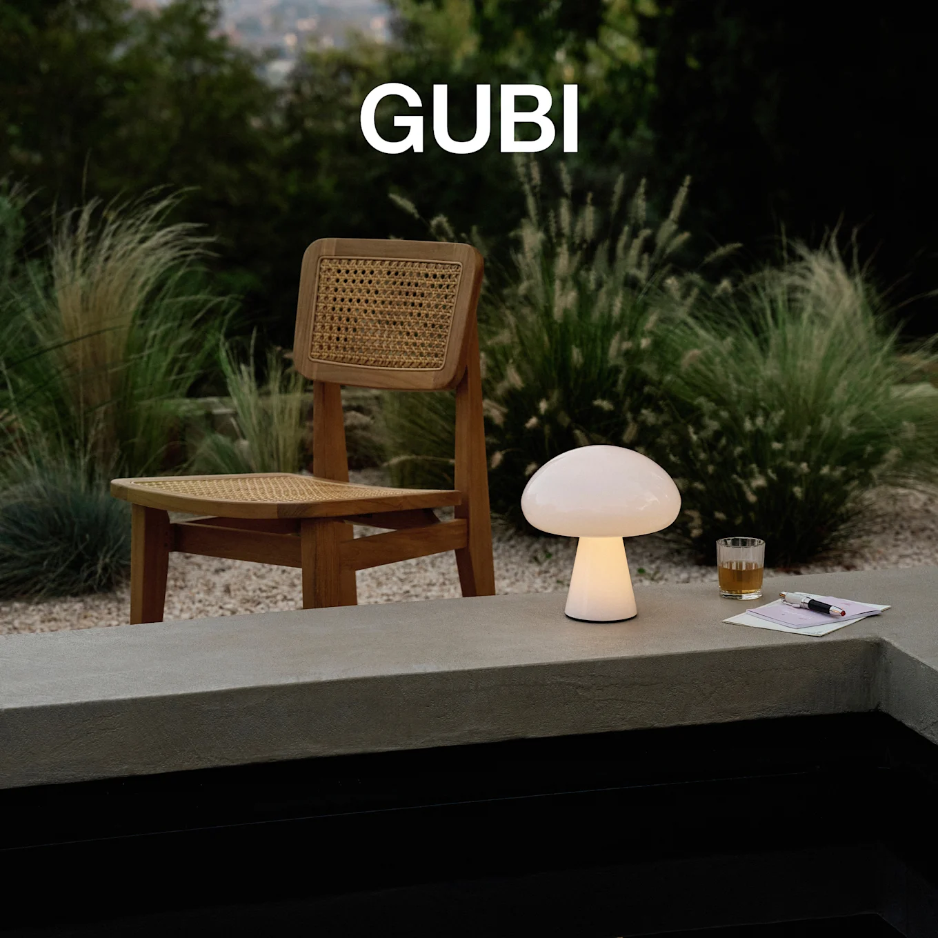 Logotipo GUBI. Um candeeiro de mesa em forma de cogumelo iluminado, um copo e uma caneta numa borda de betão, com uma cadeira de madeira e vegetação exuberante.