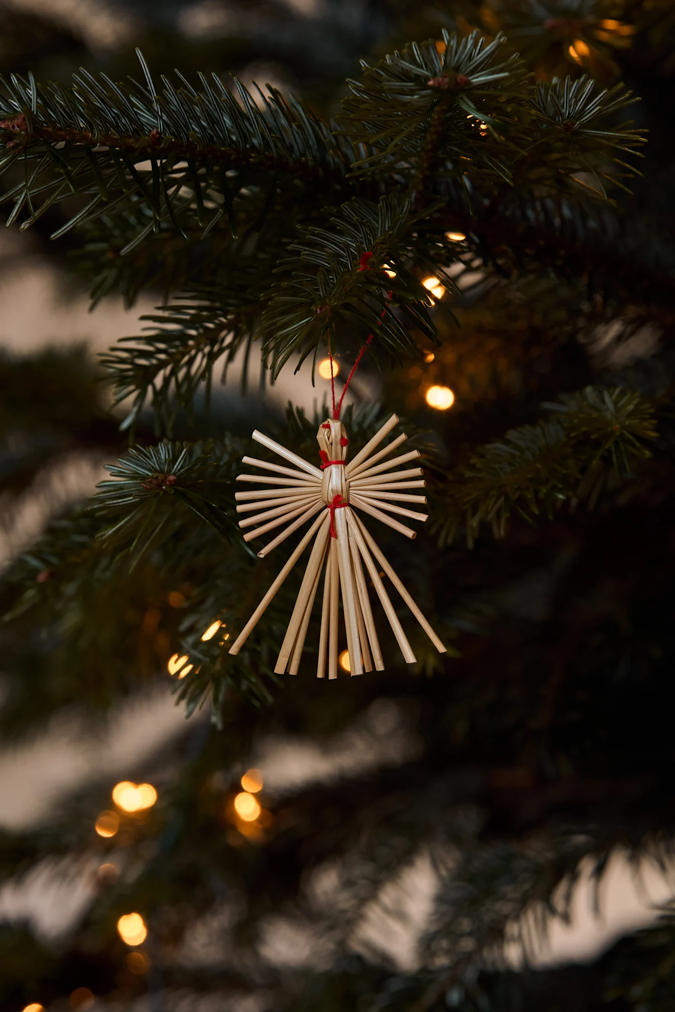 Enfeite de anjo de palha pendurado num ramo de árvore de Natal com luzes desfocadas ao fundo.