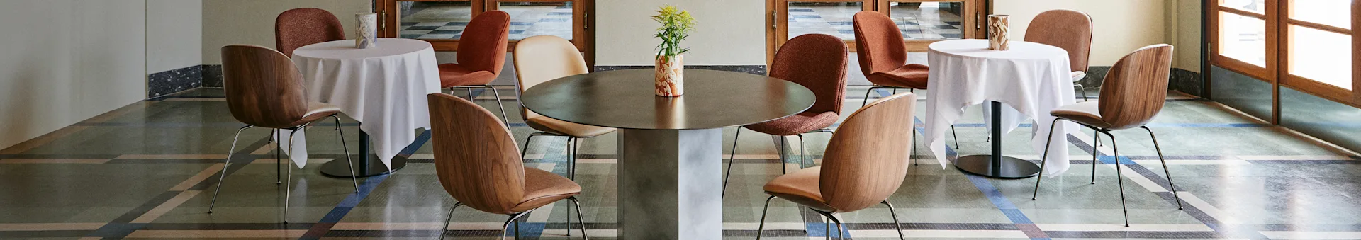 Interior de café elegante com uma grande mesa central, duas mesas menores com toalhas brancas e várias cadeiras num piso estampado.