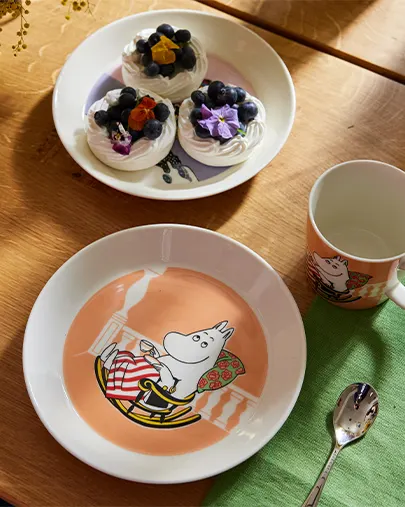 Prato, caneca e colher Moomintroll ao lado de um prato de suspiros com mirtilos e flores numa mesa de madeira.