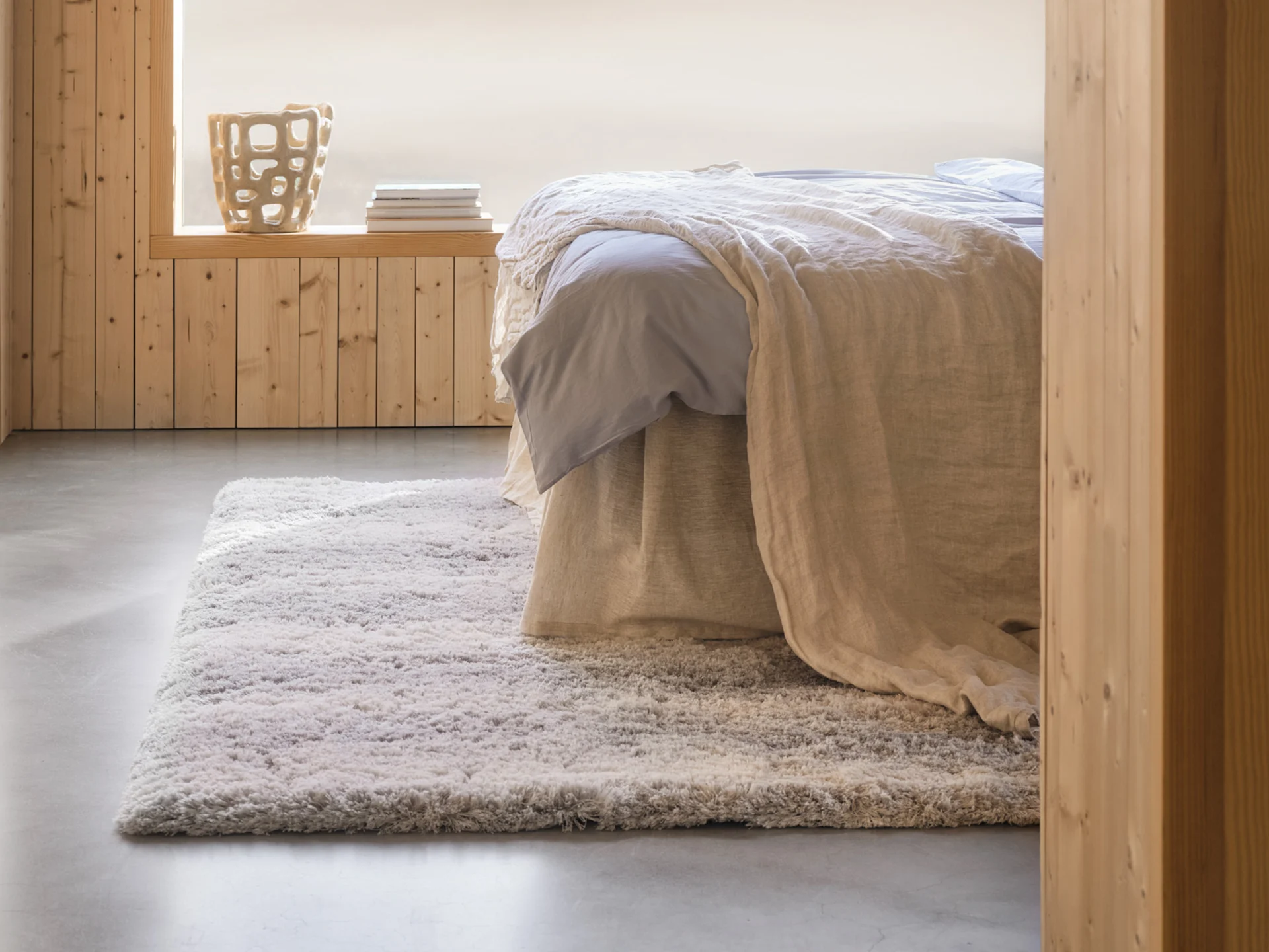 Quarto acolhedor com cama, lençóis azuis claros, manta bege, tapete felpudo, paredes de madeira e um vaso decorativo.