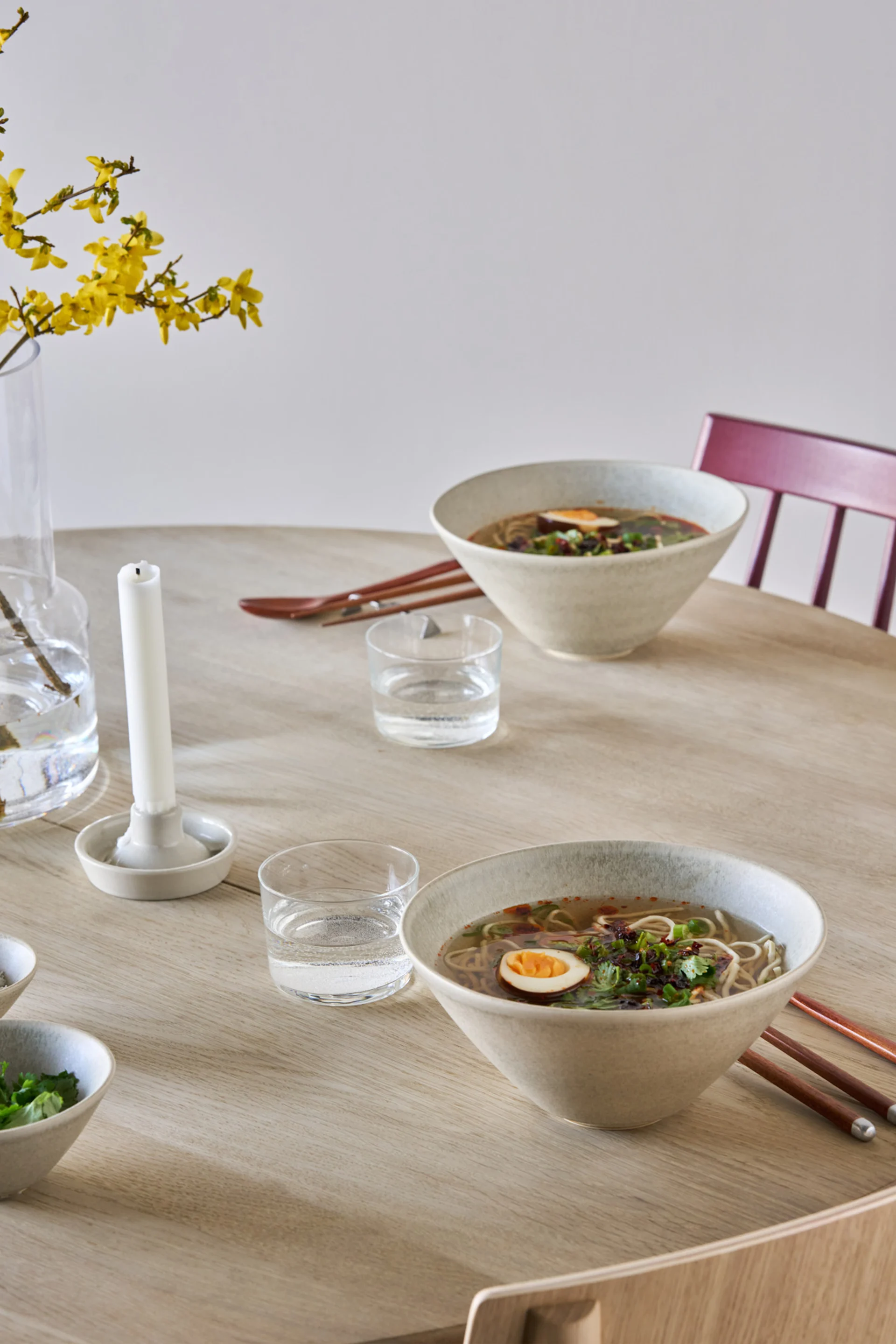 Duas tigelas de ramen com ovos e guarnição numa mesa de madeira clara, com copos de água e flores amarelas num vaso.