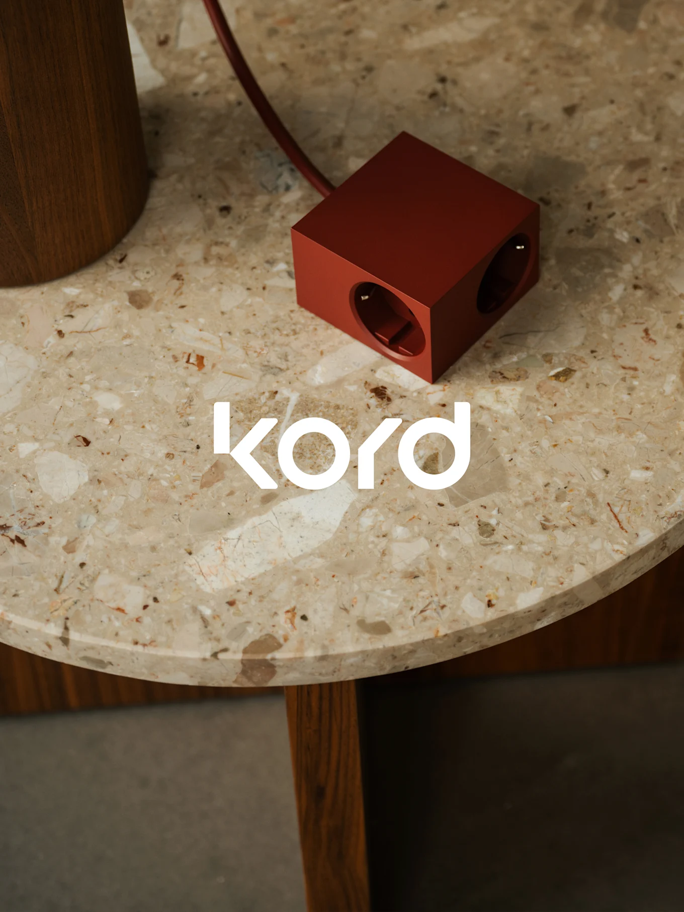 Uma régua de energia em forma de cubo, vermelho escuro, com duas tomadas visíveis, repousa sobre uma mesa redonda de mármore, com o logótipo branco "kord".