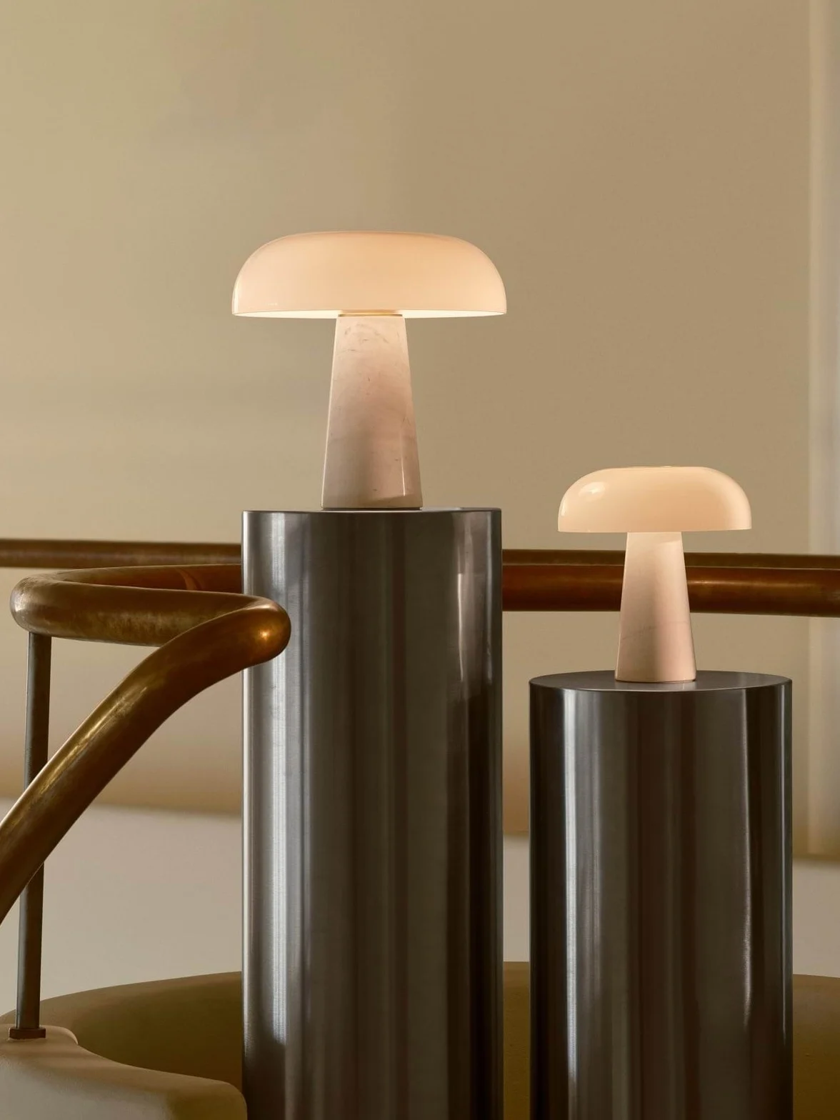 Duas lâmpadas de mesa luminosas em forma de cogumelo, uma grande e uma pequena, sobre pedestais cilíndricos ao lado de um corrimão de latão.