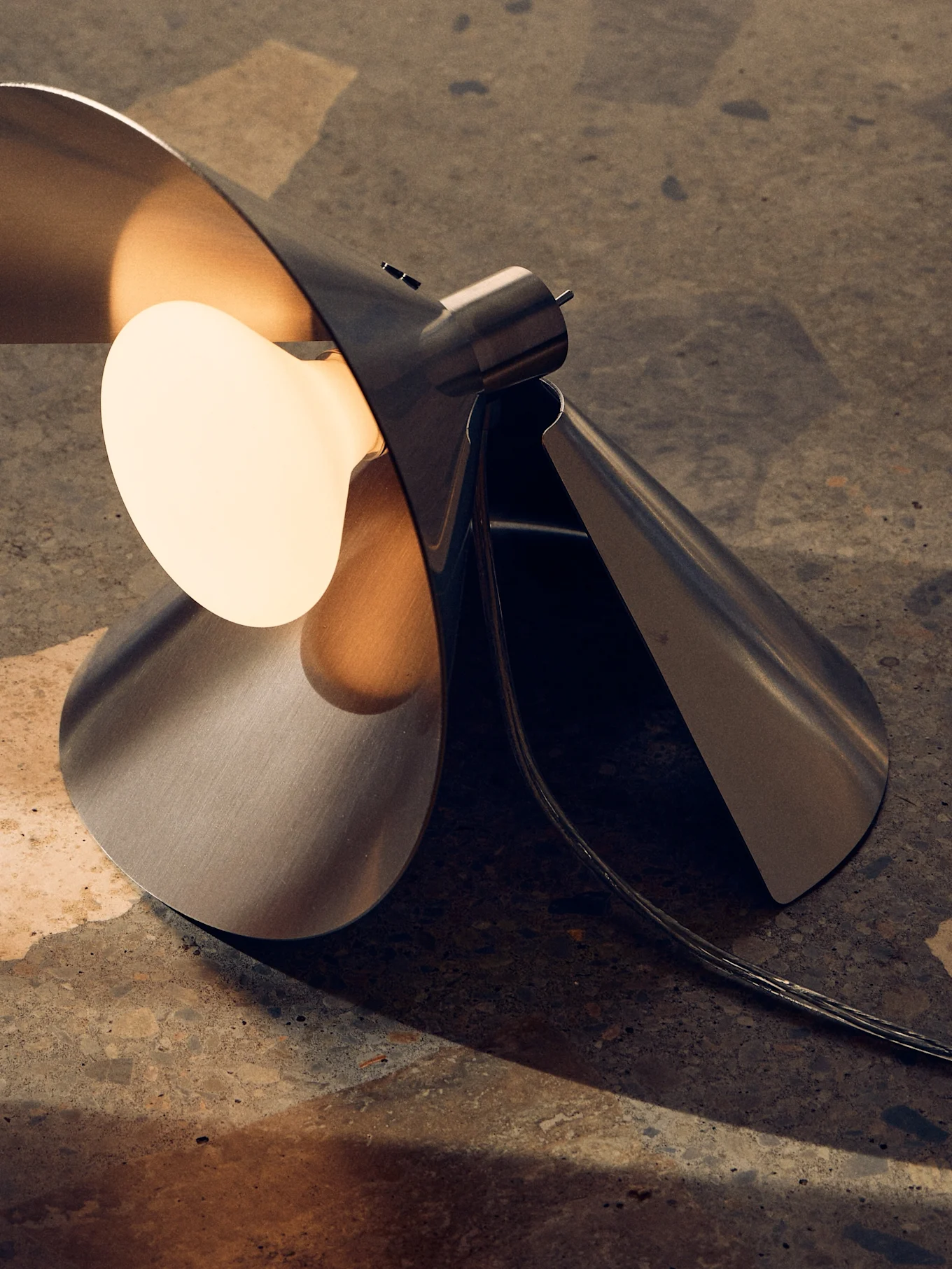 Candeeiro de mesa moderno de metal escovado com design escultural dobrado e lâmpada quente exposta em superfície texturizada.