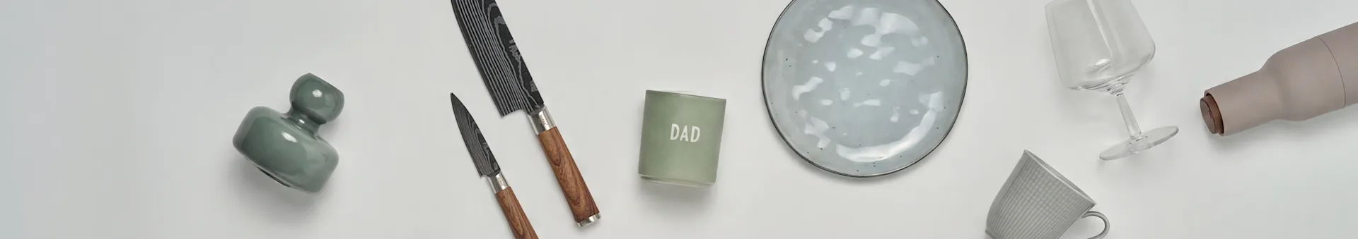 Uma vista plana de artigos de cozinha e presentes: um vaso verde, duas facas, uma vela 'DAD', um prato de cerâmica cinzento, um copo de vinho, uma garrafa bege e uma caneca cinzenta.