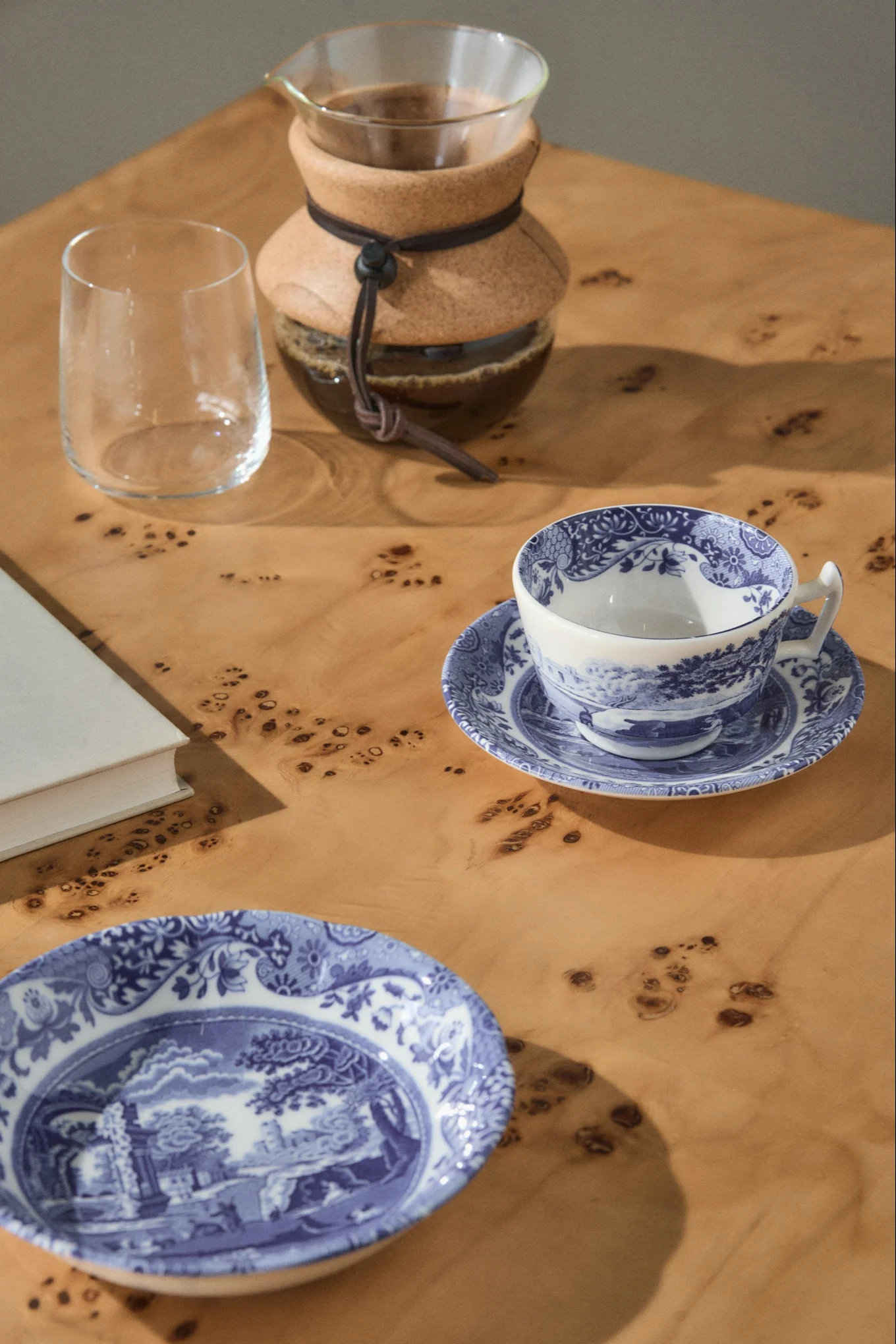 Uma cafeteira pour-over, um copo vazio, uma xícara de chá azul e branca num pires, e um prato a condizer numa mesa de madeira clara.