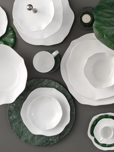 Uma coleção de louça de cerâmica branca com bordas onduladas, disposta com folhas verdes e bases de mármore sobre uma superfície escura.