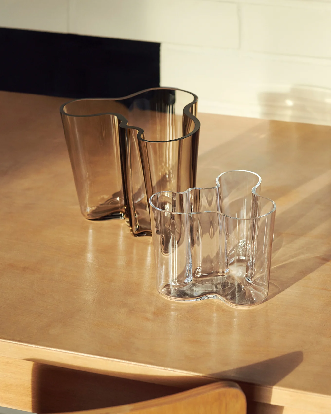 Dois vasos de vidro ondulados, um grande âmbar e um menor transparente, sobre uma superfície de madeira.