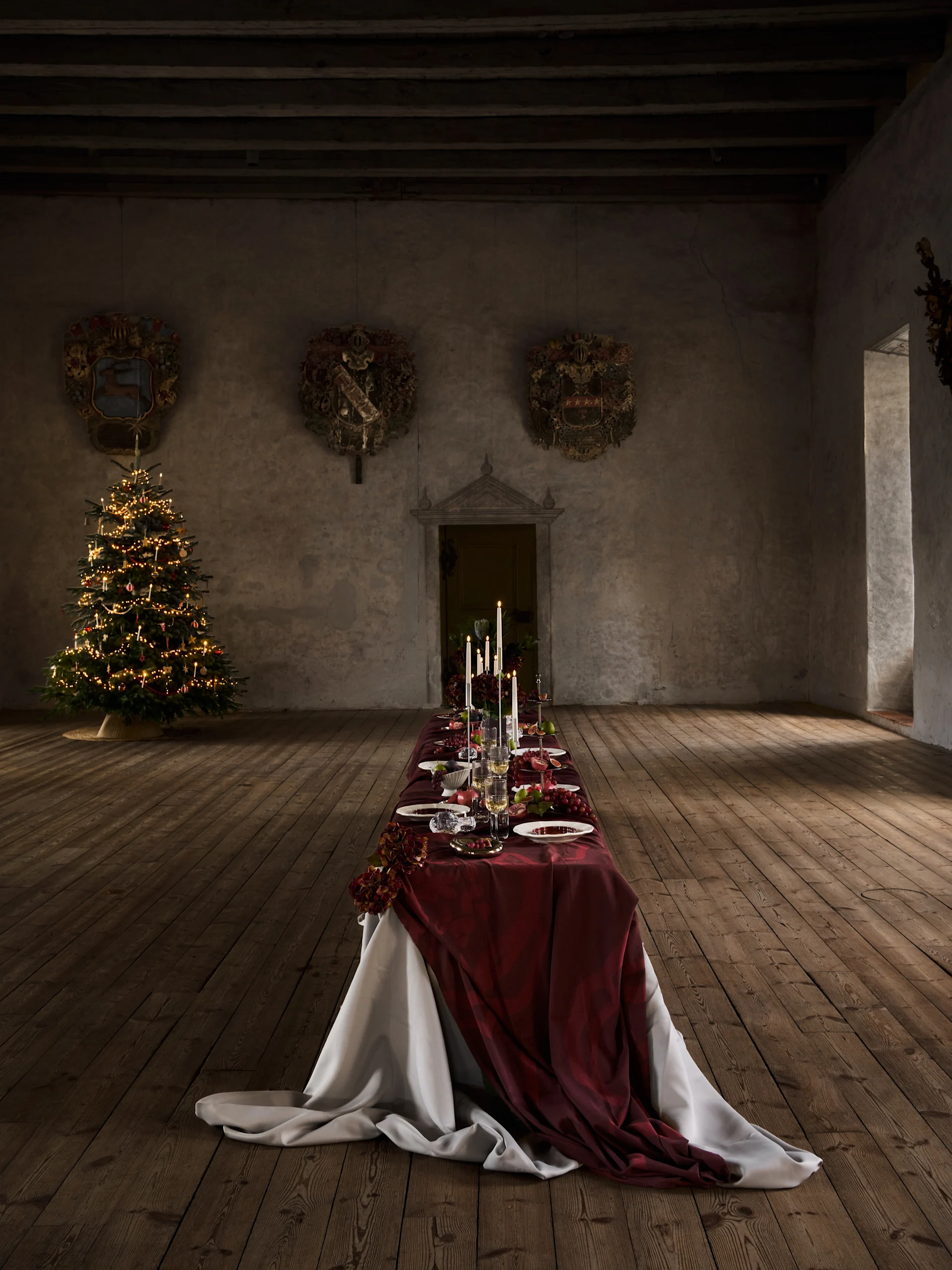 Longa mesa de jantar de Natal com toalha vermelha, velas, frutas e talheres, ao lado de uma árvore de Natal iluminada em um salão antigo.