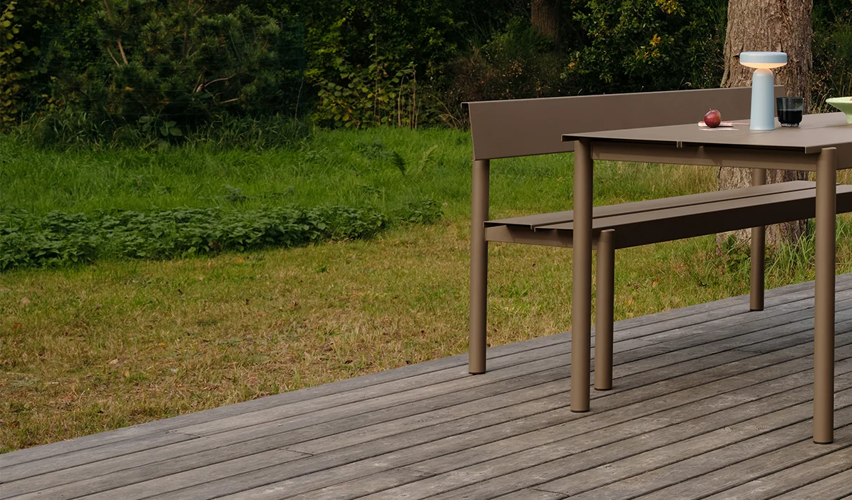 Mesa e banco de jantar modernos em taupe num deck de madeira, com um candeeiro portátil e maçãs na mesa.
