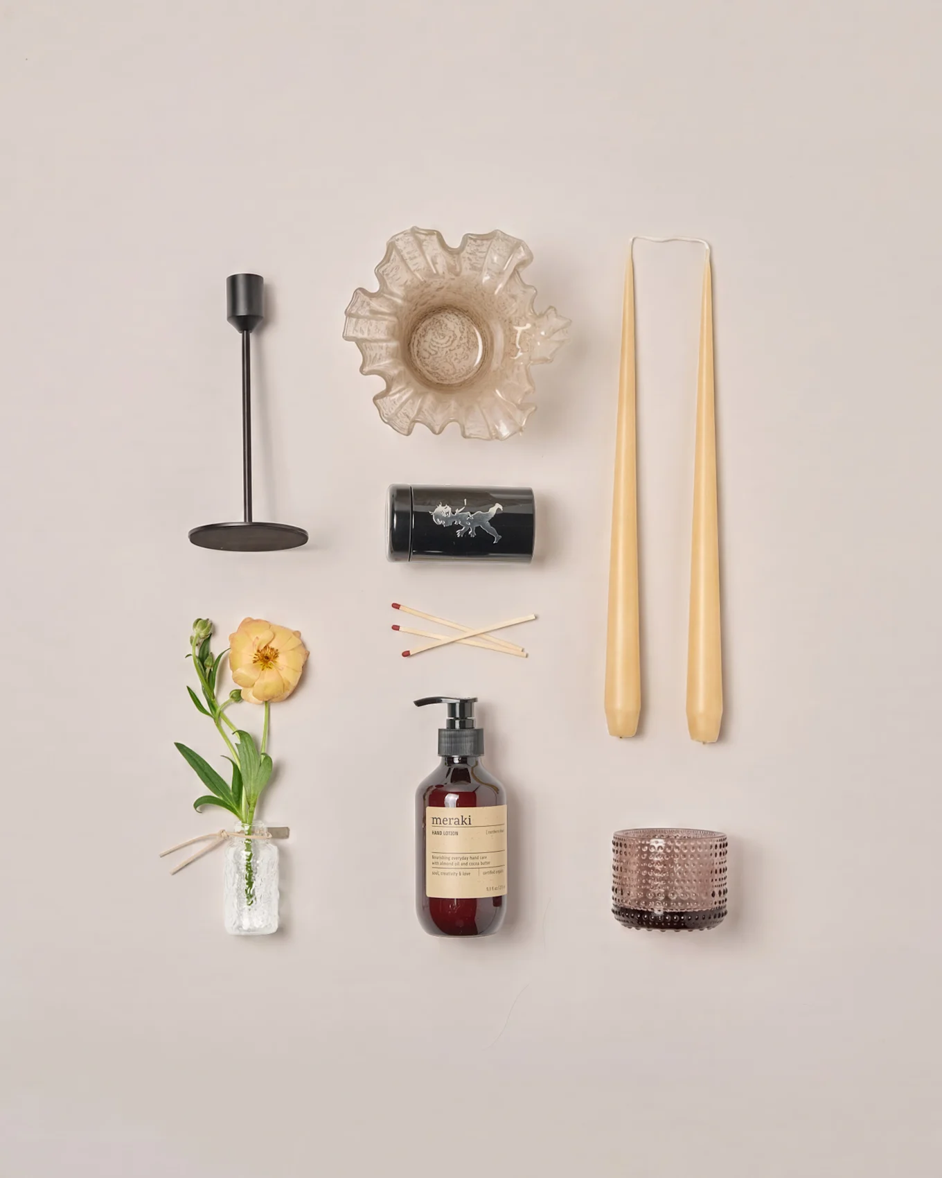 Arranjo plano de vários itens de decoração para casa: castiçal, tigela com babados, velas cônicas, fósforos, loção, porta-velas e uma flor de ranúnculo.