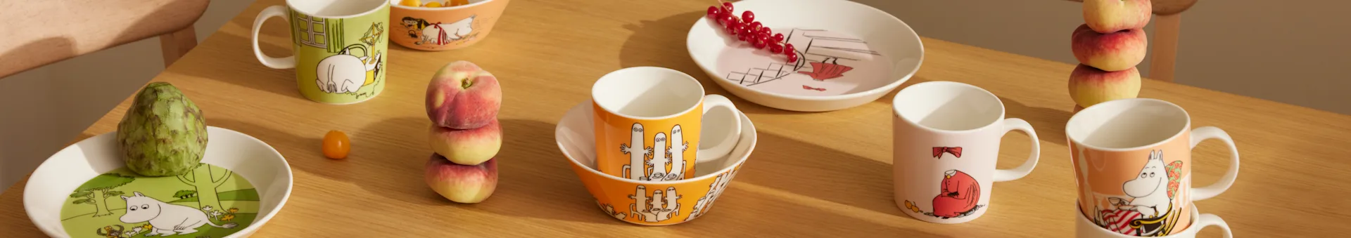 Louça Moomin em cores e designs variados, com canecas, tigelas e pratos, e fruta fresca numa mesa de madeira.