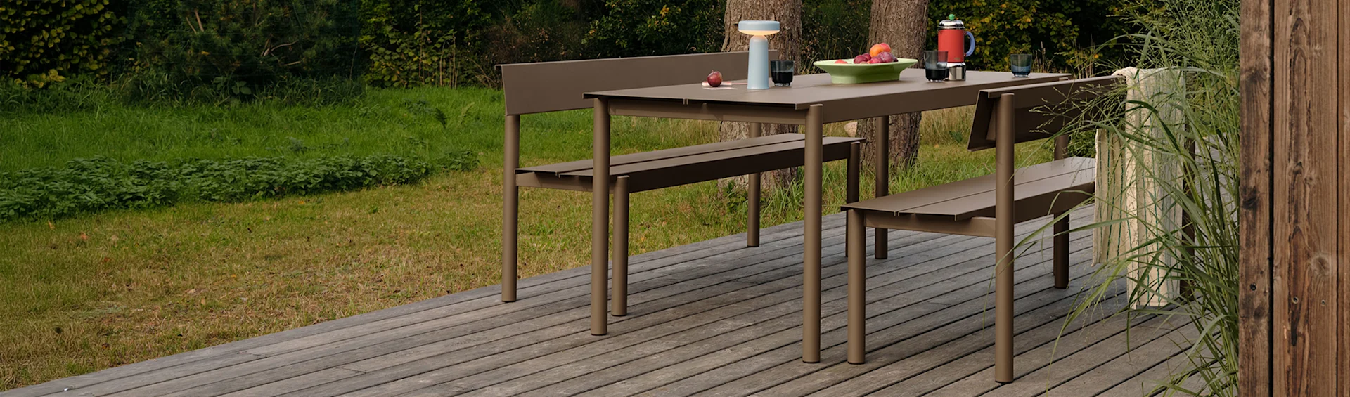 Moderno conjunto jantar exterior taupe: mesa, dois bancos, fruta, candeeiro, bule de café em deque de madeira, verde.