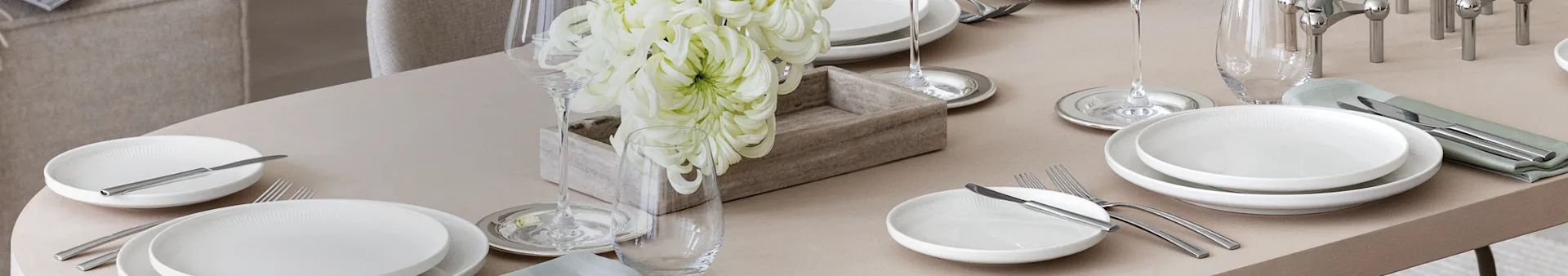 Uma mesa de jantar elegantemente posta com pratos brancos, talheres de prata, copos de vinho e água, e um centro de mesa com grandes flores brancas numa caixa de madeira.