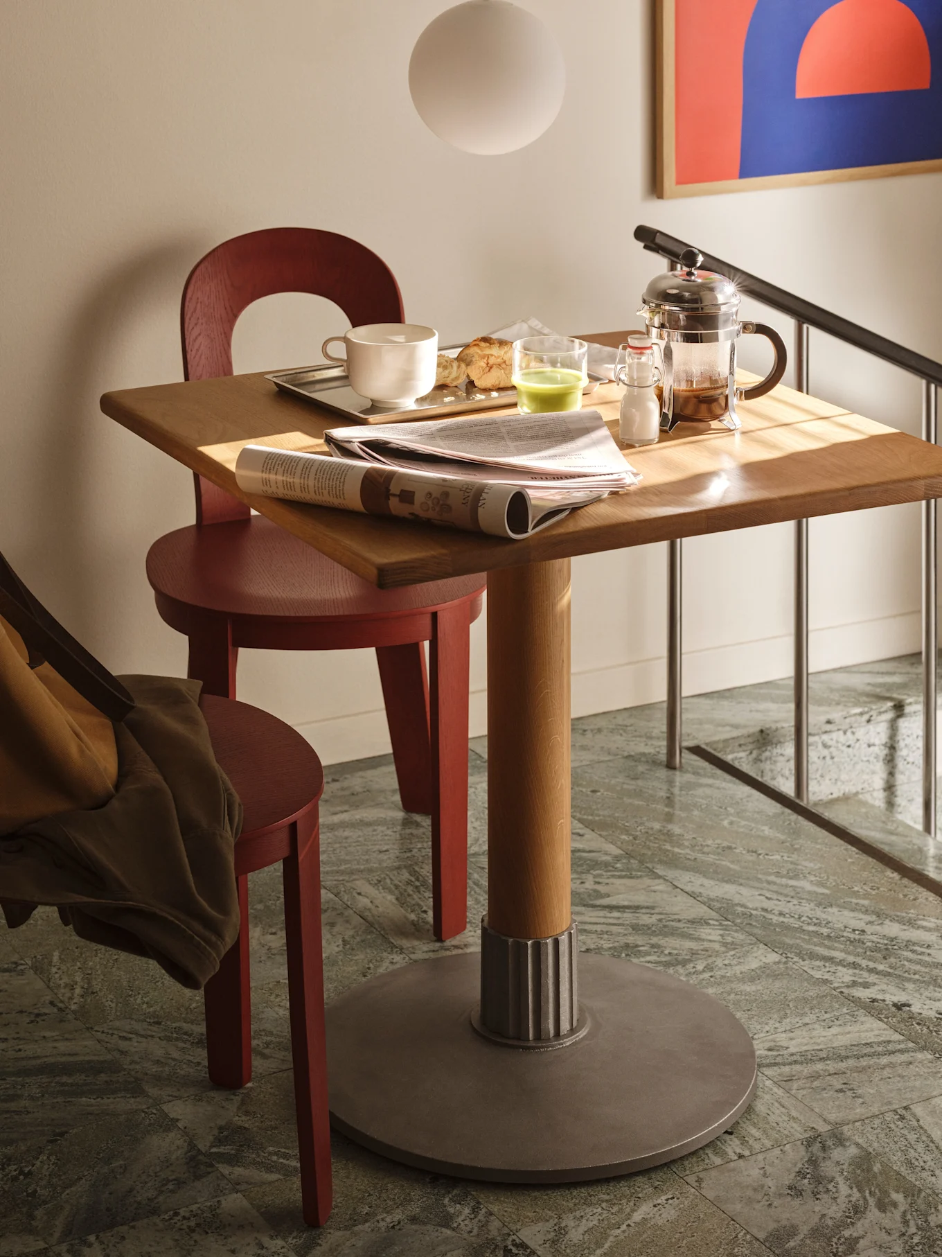 Uma mesa quadrada de madeira com café da manhã, jornal e prensa francesa, flanqueada por duas cadeiras vermelhas em piso.